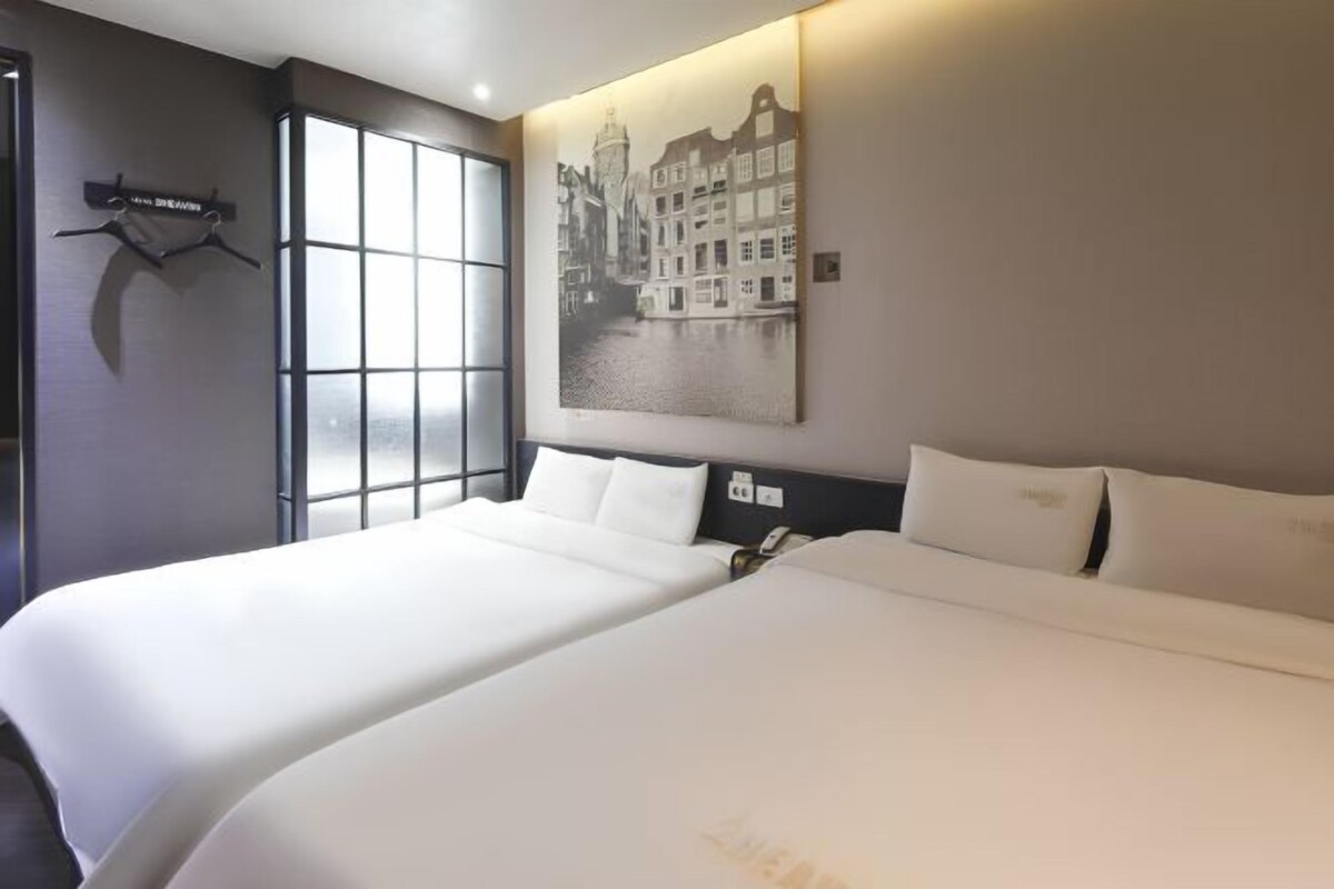 A hotel room with two beds, a large window, and a cityscape painting on the wall.