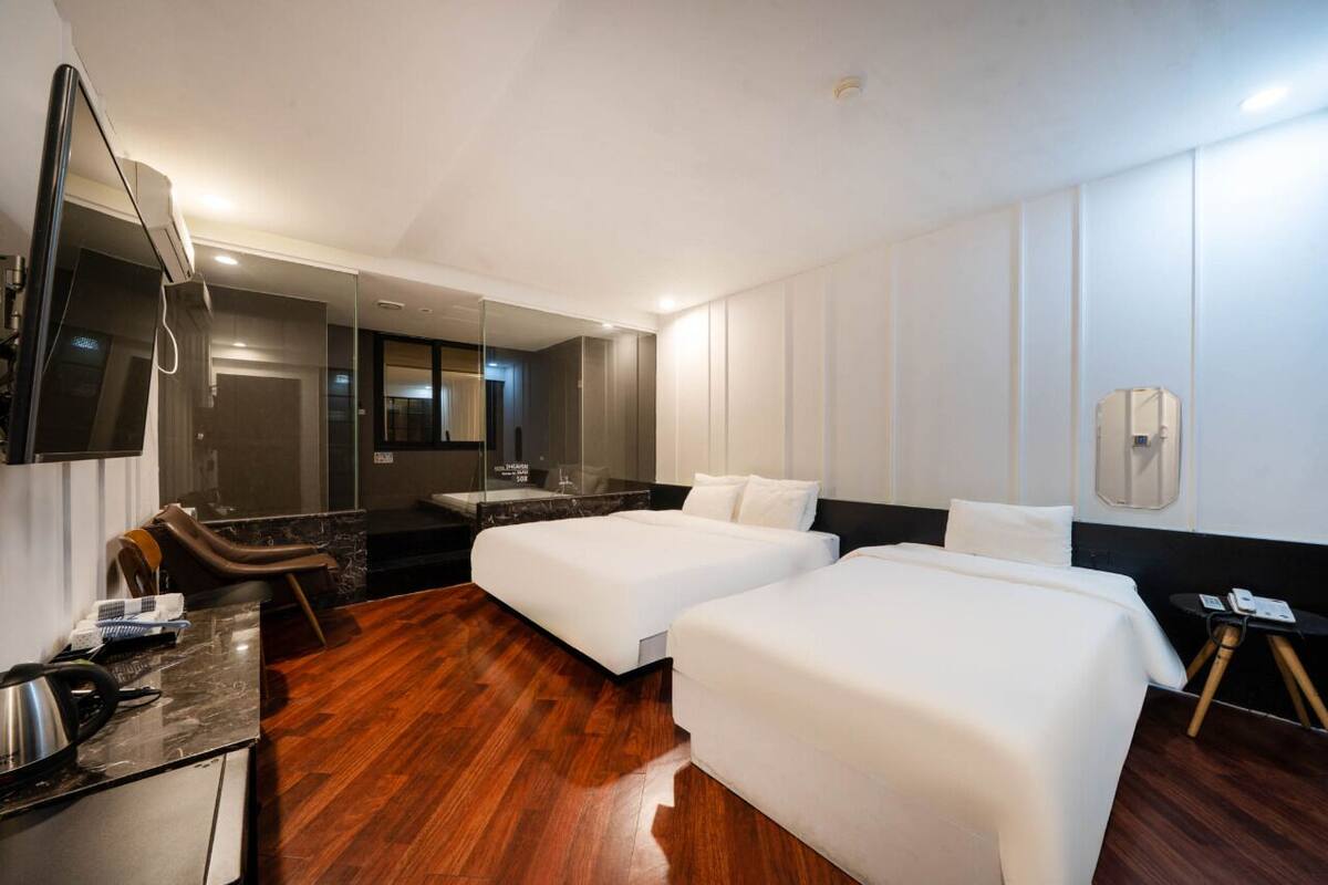 A hotel room with two beds, a desk with a chair, a mirror, and a bathroom visible through a glass door.