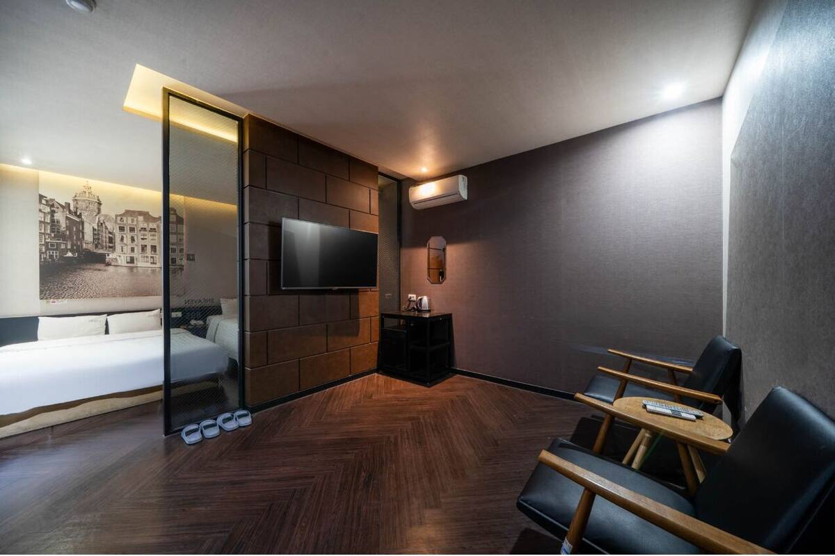 A modern hotel room with a bed, a TV, a seating area with a table, and a shoe rack.