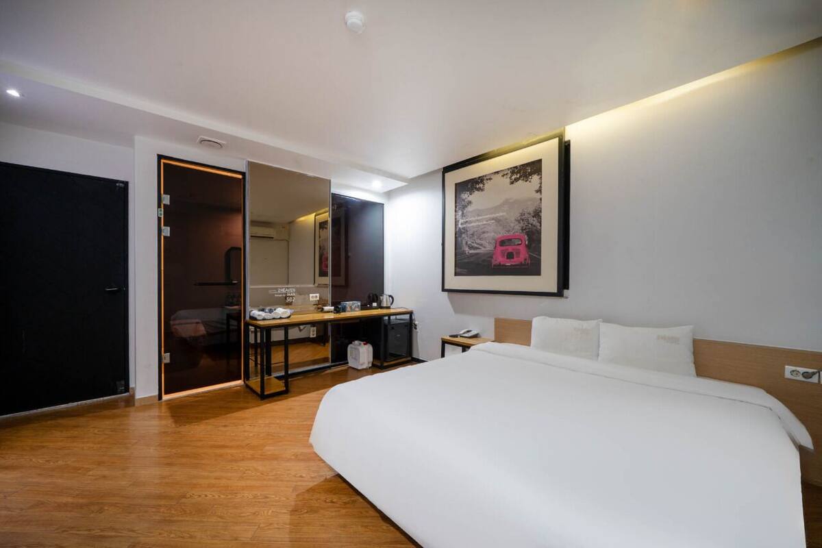 A hotel room with a large bed, a desk with a chair, a mirror, and a painting on the wall.