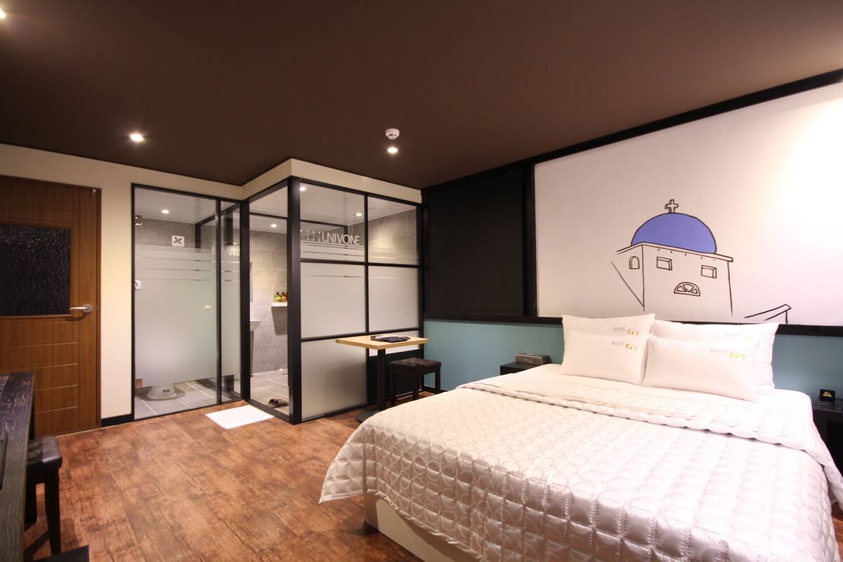 Ein modernes Hotelzimmer mit einem großen Bett, einem Schreibtisch und einer Glaskabine, die das Badezimmer abtrennt.