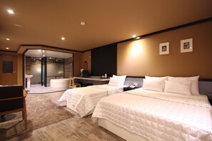 Habitación de hotel con dos camas, un escritorio, una silla, baño y zona de ducha con paredes de vidrio.
