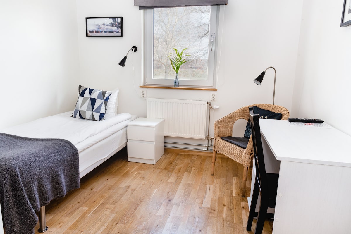 A compact bedroom with a bed, bedside table, a small desk with a chair, a window with a plant, and a radiator.