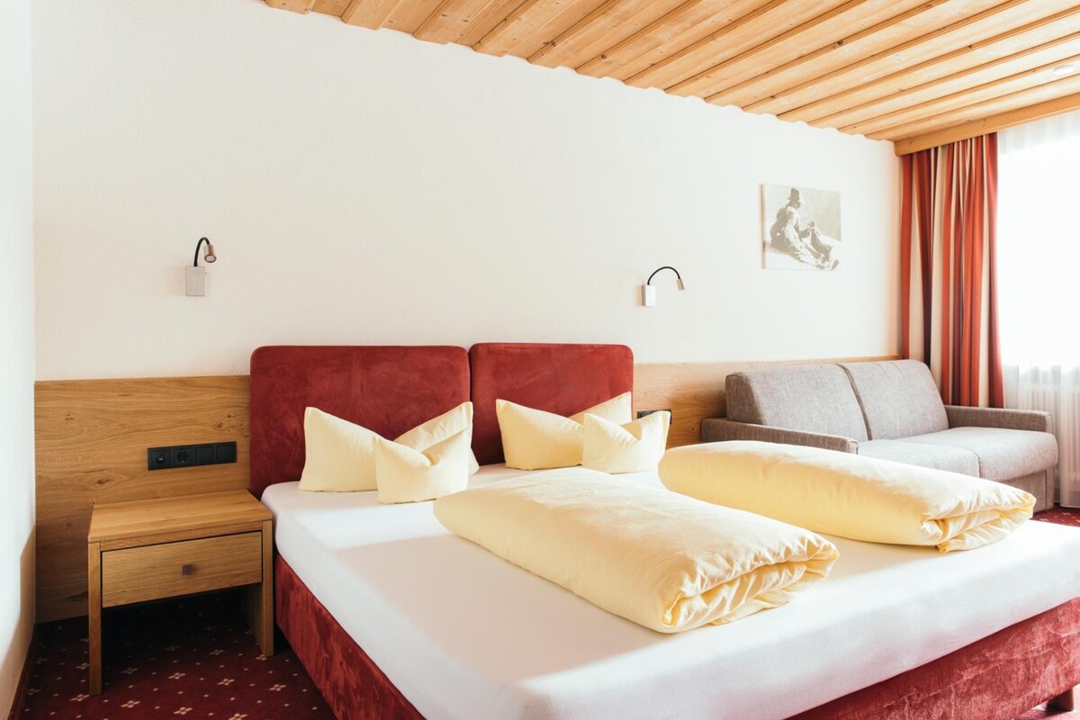 A hotel room with a wooden headboard, a bed with white linens and multiple pillows, a grey sofa, a small wooden nightstand, and two wall-mounted light fixtures.