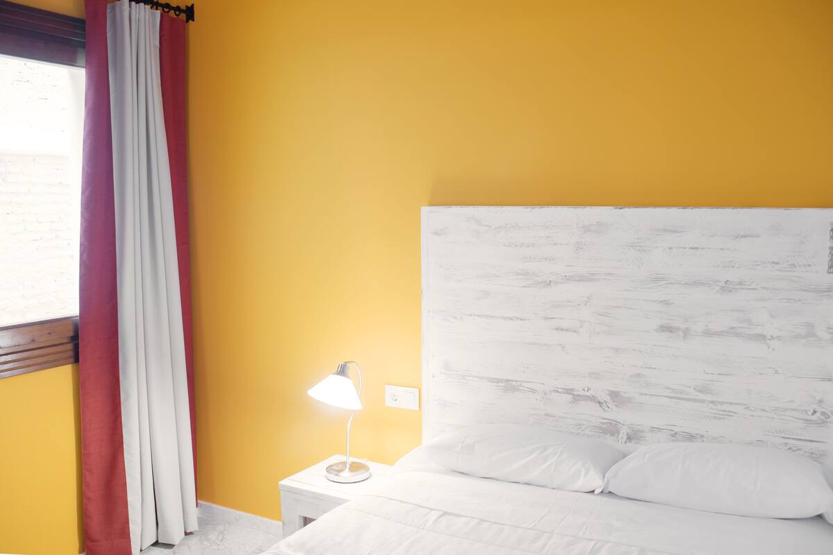 A room with a yellow wall, white curtains, a white lamp on a bedside table, and a white bed with pillows.