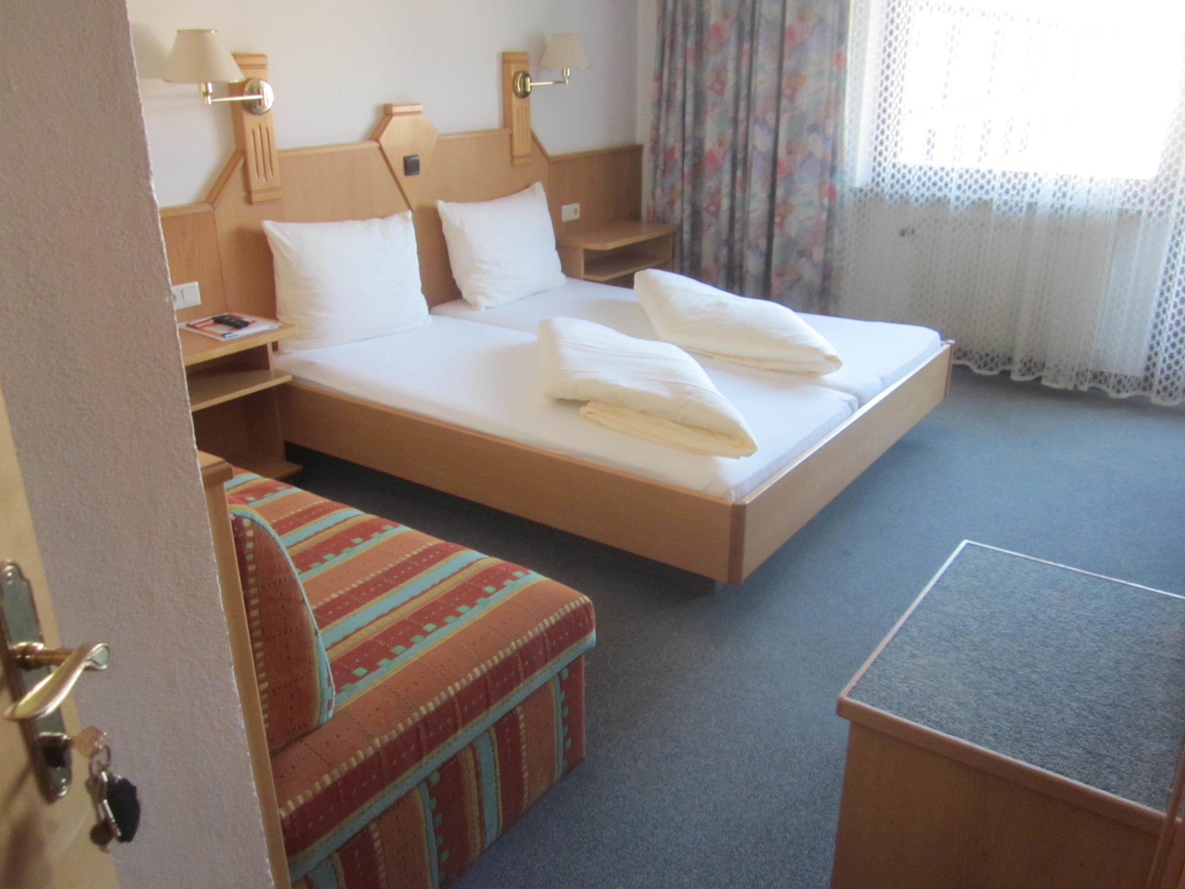 A hotel room with a large bed, two bedside tables, a small desk, a colorful bench, and a window with curtains.