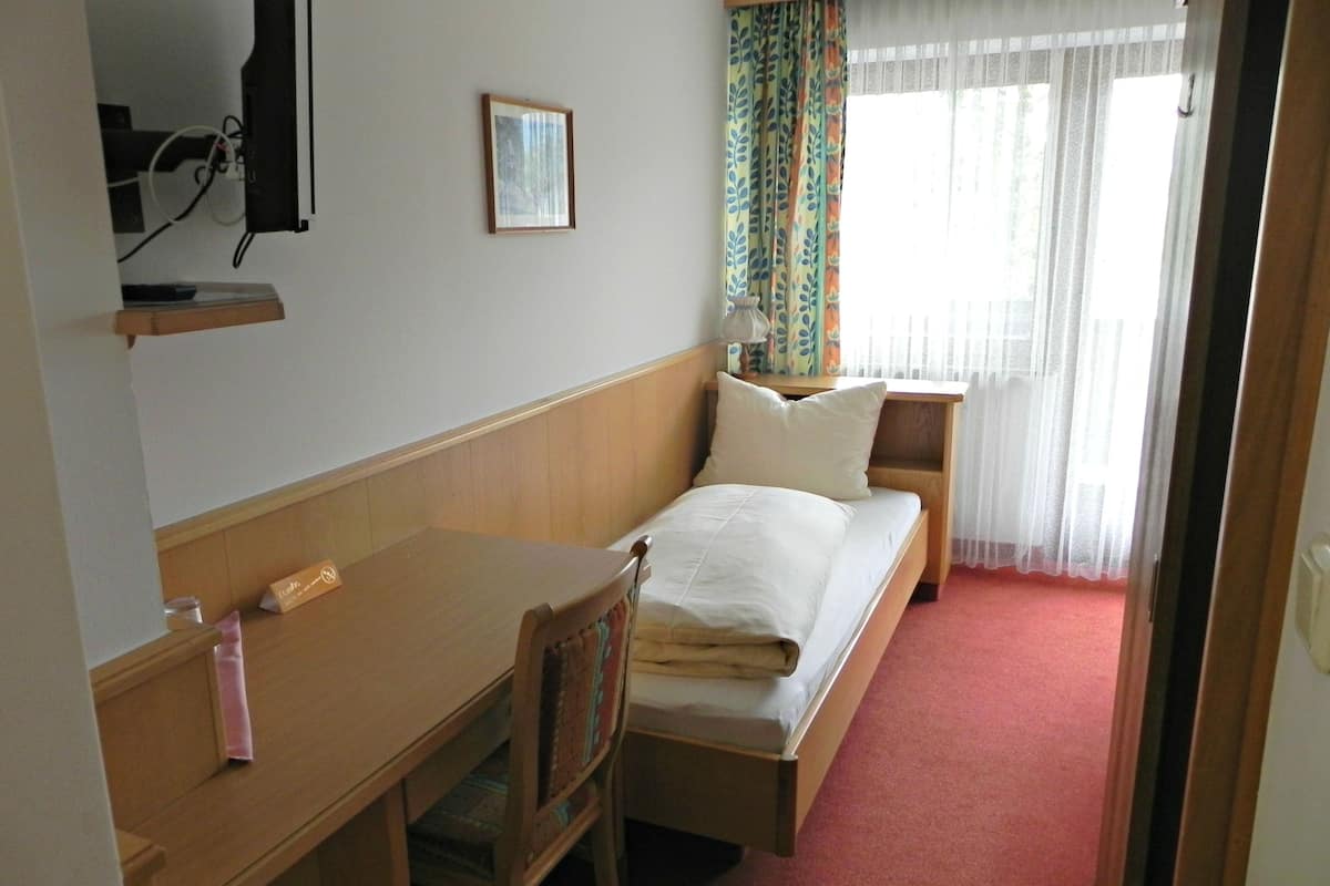 Habitació d'hotel amb un llit individual, un escriptori de fusta, una cadira, una tauleta de nit, un televisor fixat a la paret i una finestra amb cortines.