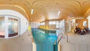 A modern indoor swimming pool with a wooden ceiling, clear glass walls, and a seating area with wicker chairs. - Hotel Sonnhof (Radfeld)