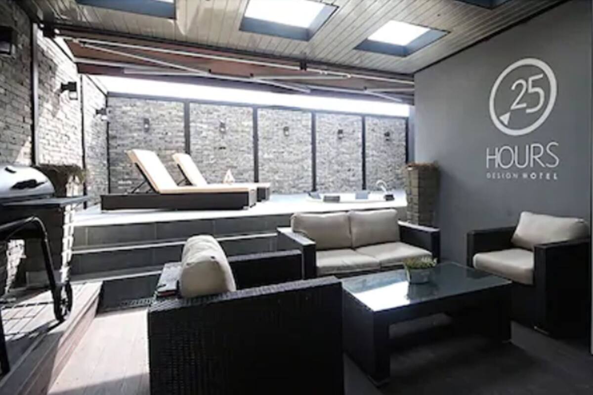 Un moderno vestíbulo de hotel con una pared de piedra, una sala de estar con sofás y una mesa de café, y una pared de cristal con el texto "25 HOURS BELTON HOTEL".