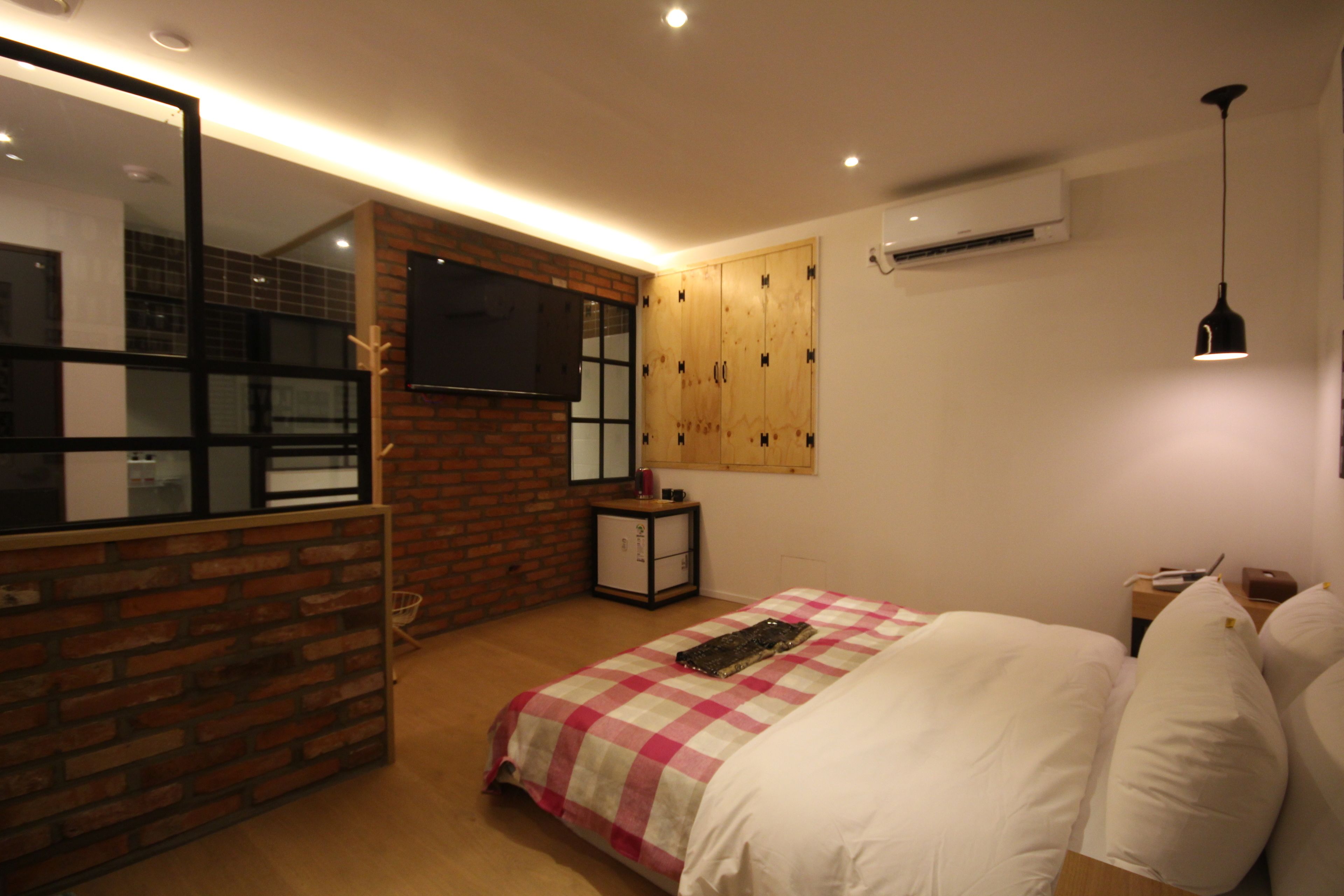 A bedroom with a checkered bedspread, a television mounted on a brick wall, and a wooden wardrobe.