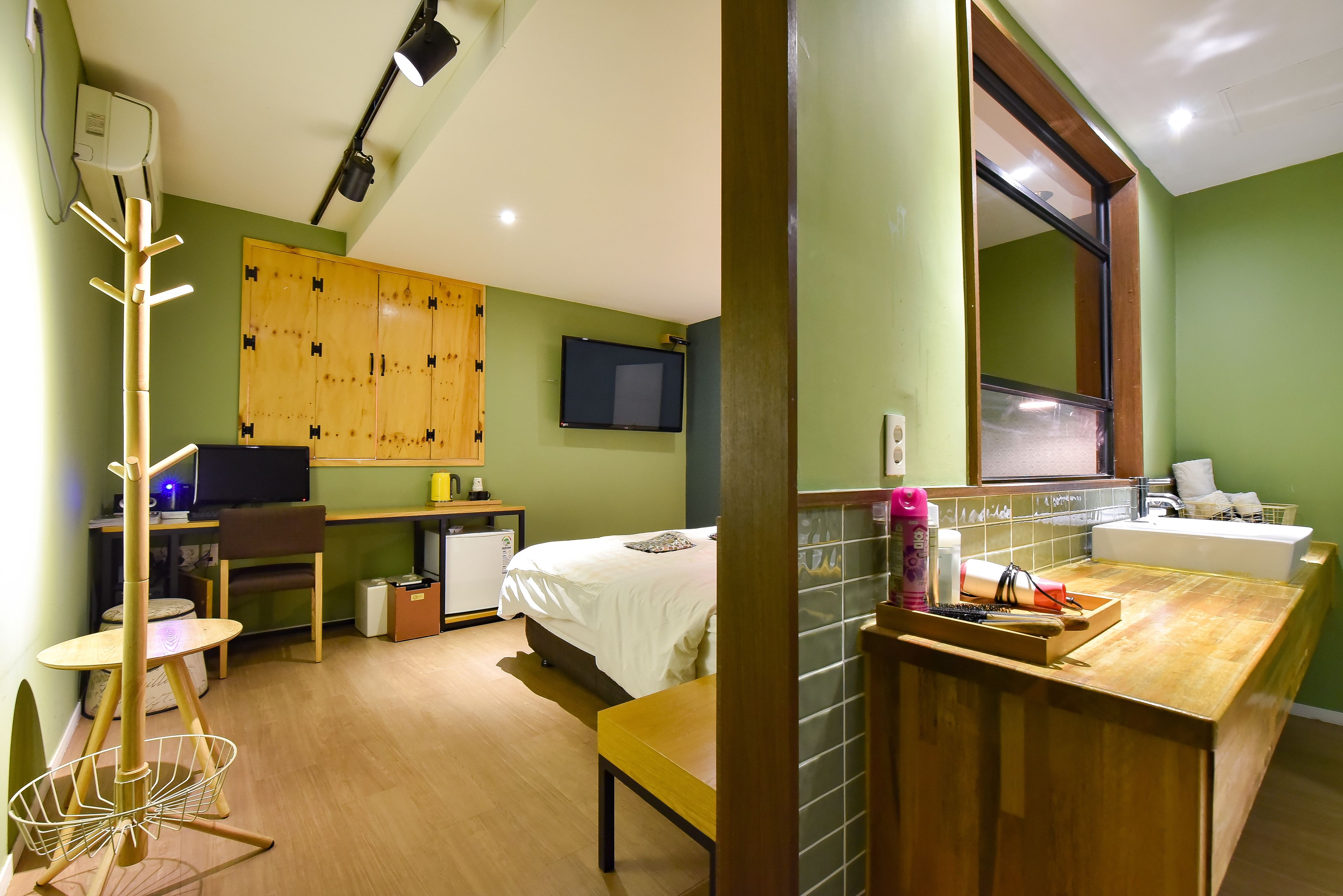 Ein kompaktes Hotelzimmer mit Bett, Schreibtisch inklusive Computer, einer kleinen Küchenzeile und einem Badezimmer mit Waschbecken und Spiegel.