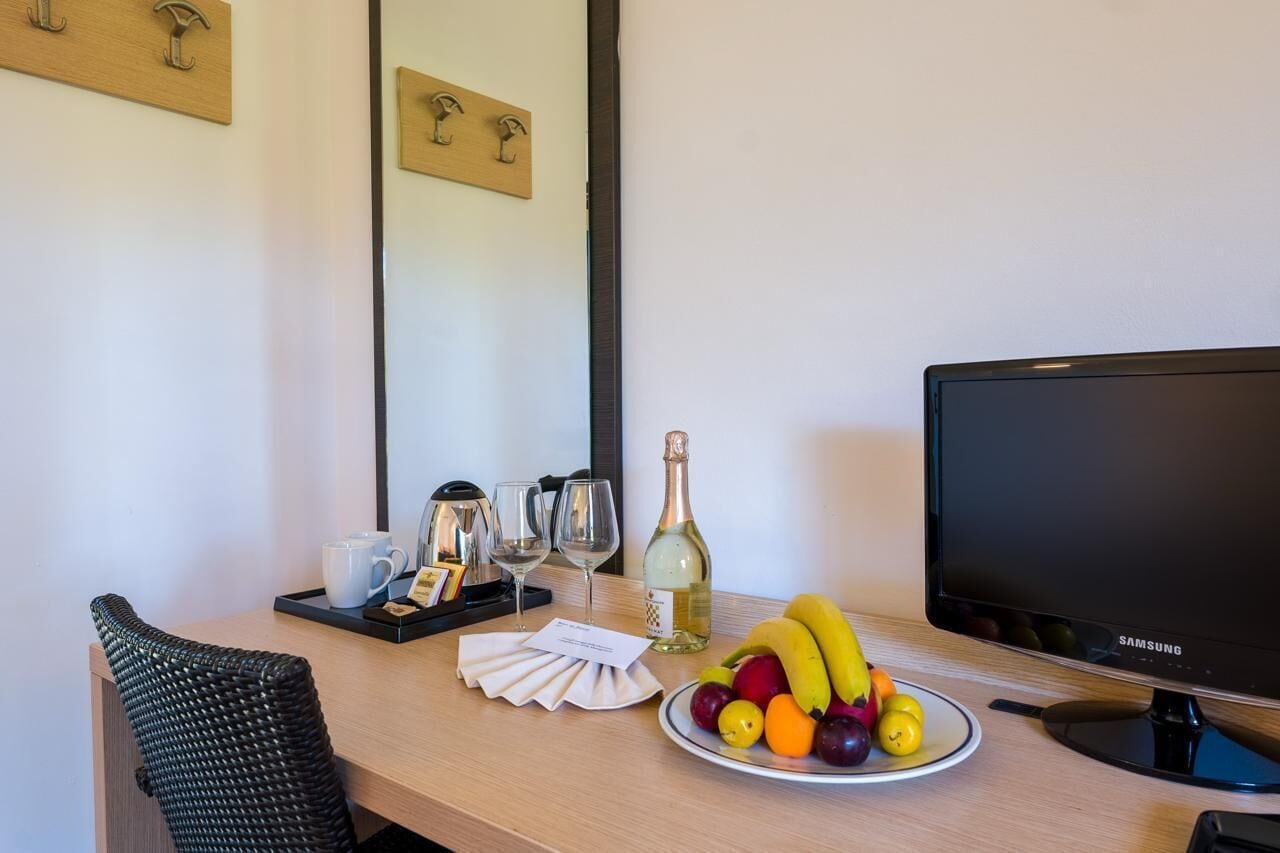 Une chambre d’hôtel avec un bureau en bois, une télévision, une bouteille de vin, des verres, une assiette de fruits et un fauteuil en osier noir.