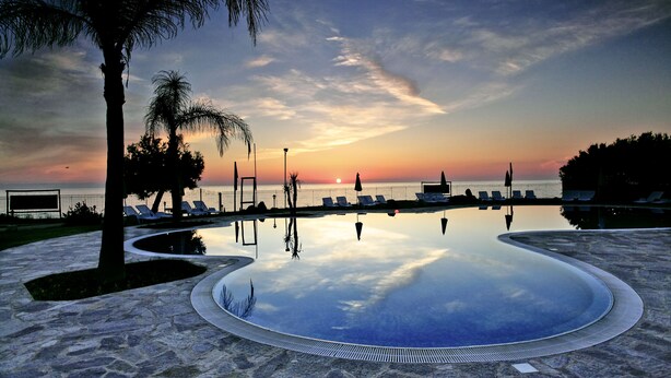 A pool with a curved edge, surrounded by lounge chairs and palm trees, with a sunset in the background. - Borgo del Principe by Valentour (Zambrone)