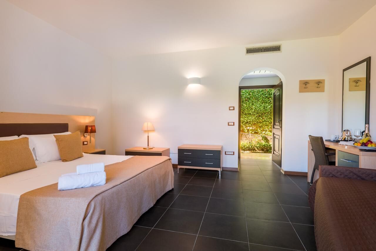 A hotel room with a bed, bedside tables, a desk, and a view of greenery through an archway.