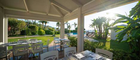 An outdoor dining area with tables and chairs, a pool, and lush greenery in the background.