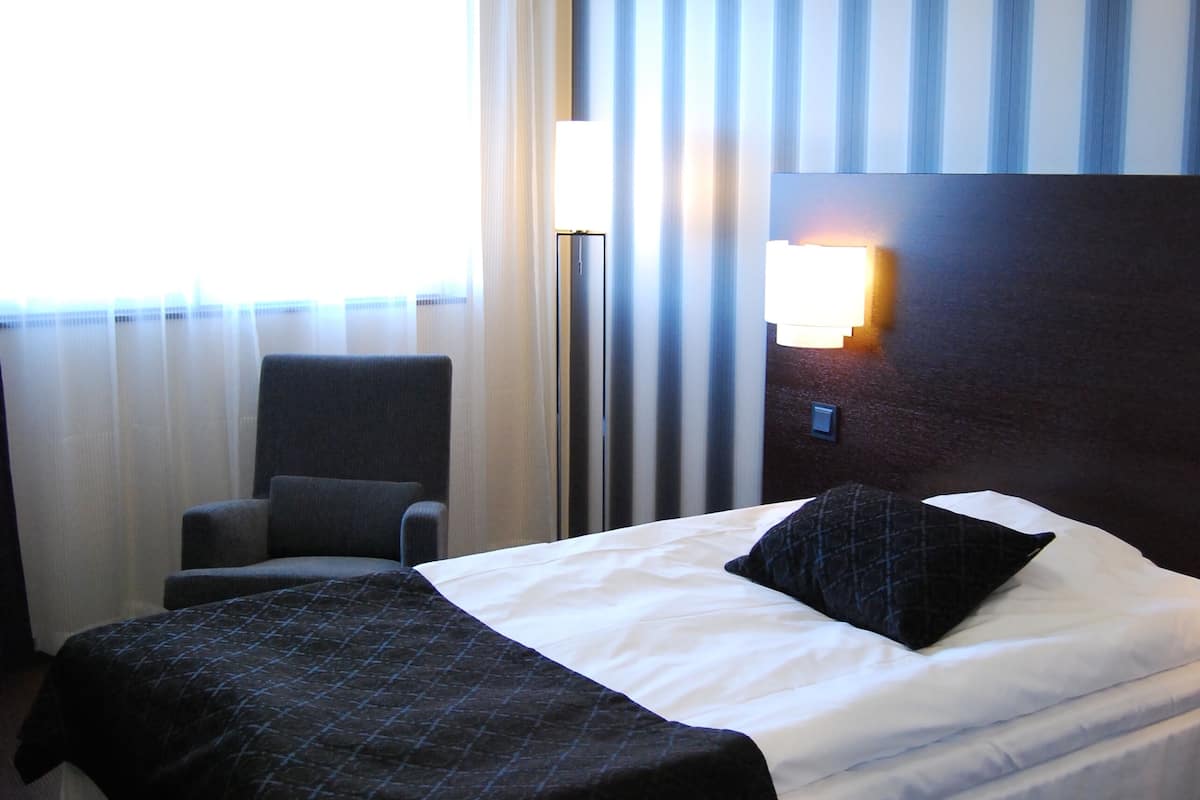 Une chambre d’hôtel avec un grand lit, une chaise, une fenêtre avec des rideaux et un luminaire fixé au mur.