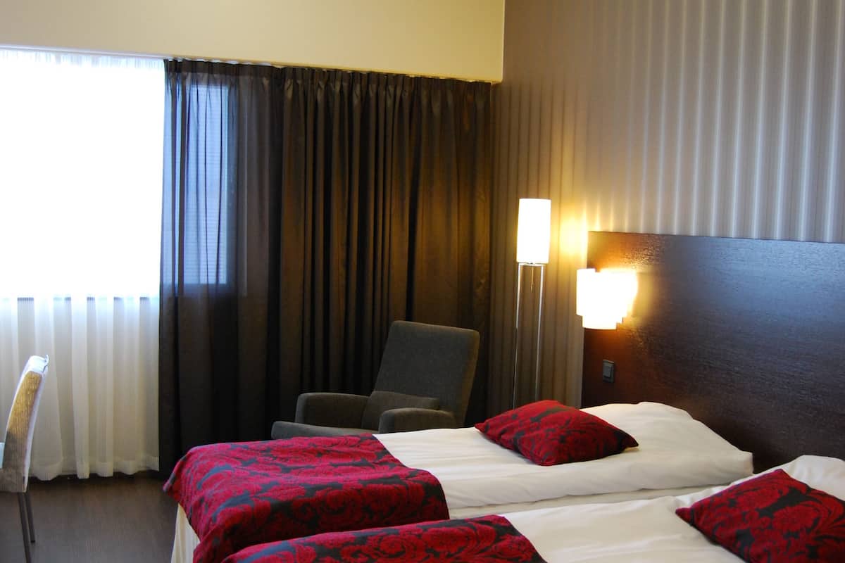 Une chambre d’hôtel avec deux lits, une chaise, une lampe et une fenêtre avec des rideaux.