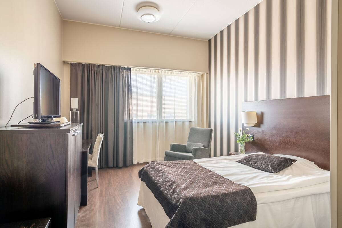Une chambre d’hôtel avec un lit, un bureau avec une télévision, une chaise et une fenêtre avec des rideaux.