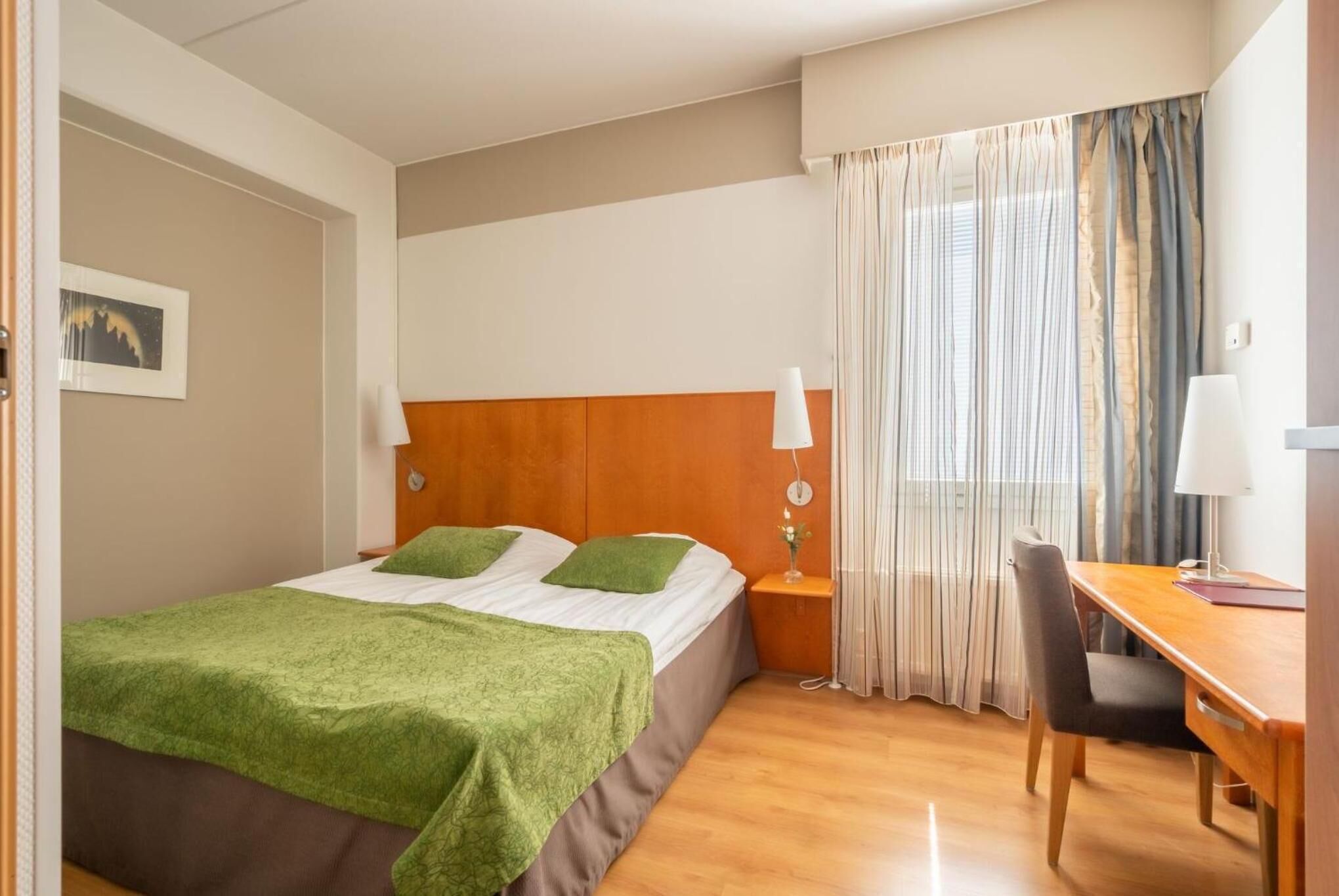 Ein Hotelzimmer mit Bett, Schreibtisch, Stuhl, Nachttisch und einem Fenster mit Vorhängen.