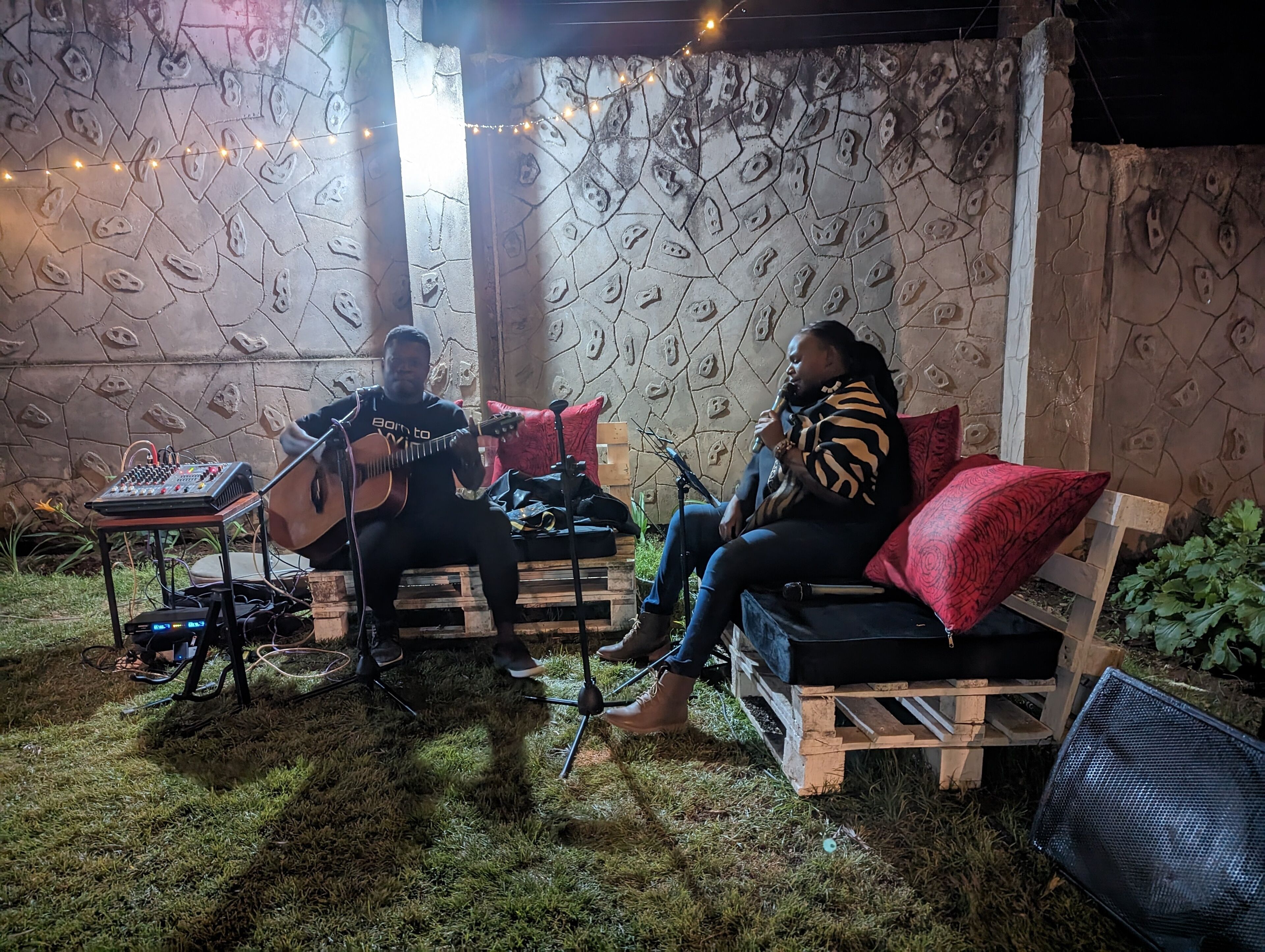 Two people seated on a wooden bench outdoors, one playing an acoustic guitar and the other singing into a microphone.