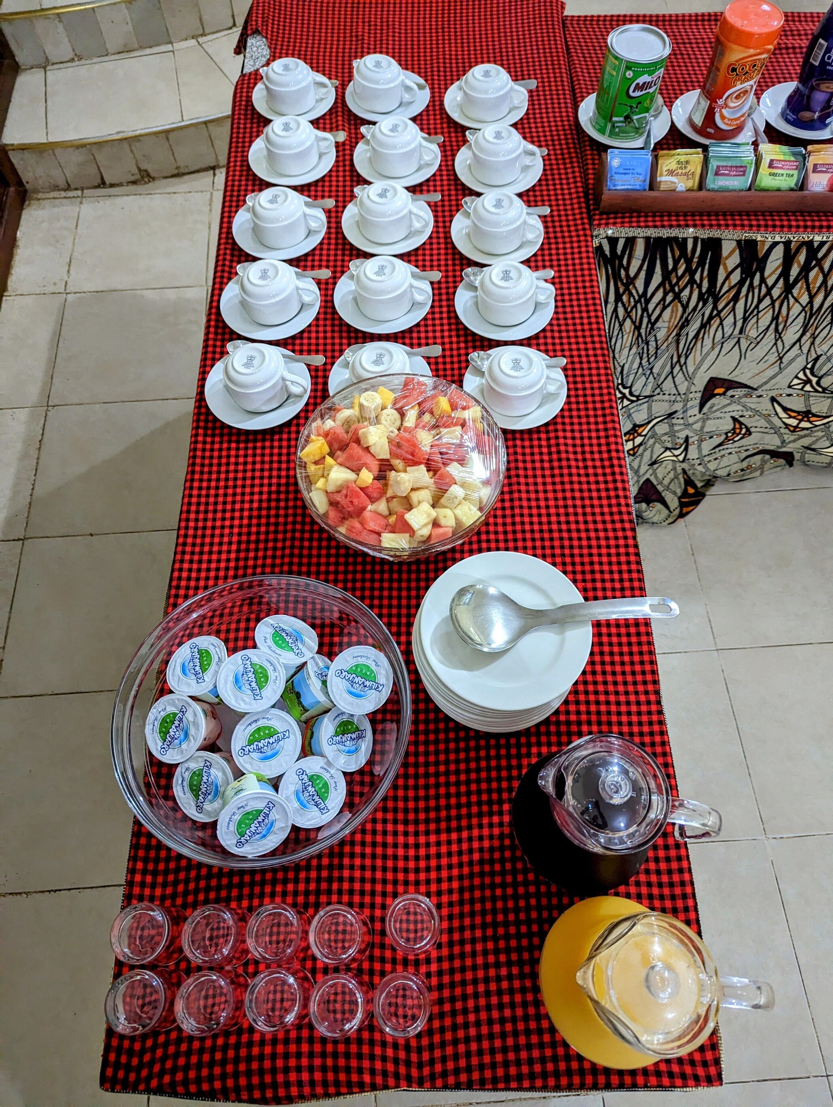 En buffetbod med hvide kopper, en skål med frugt, yoghurtbeholdere og en kande juice.
