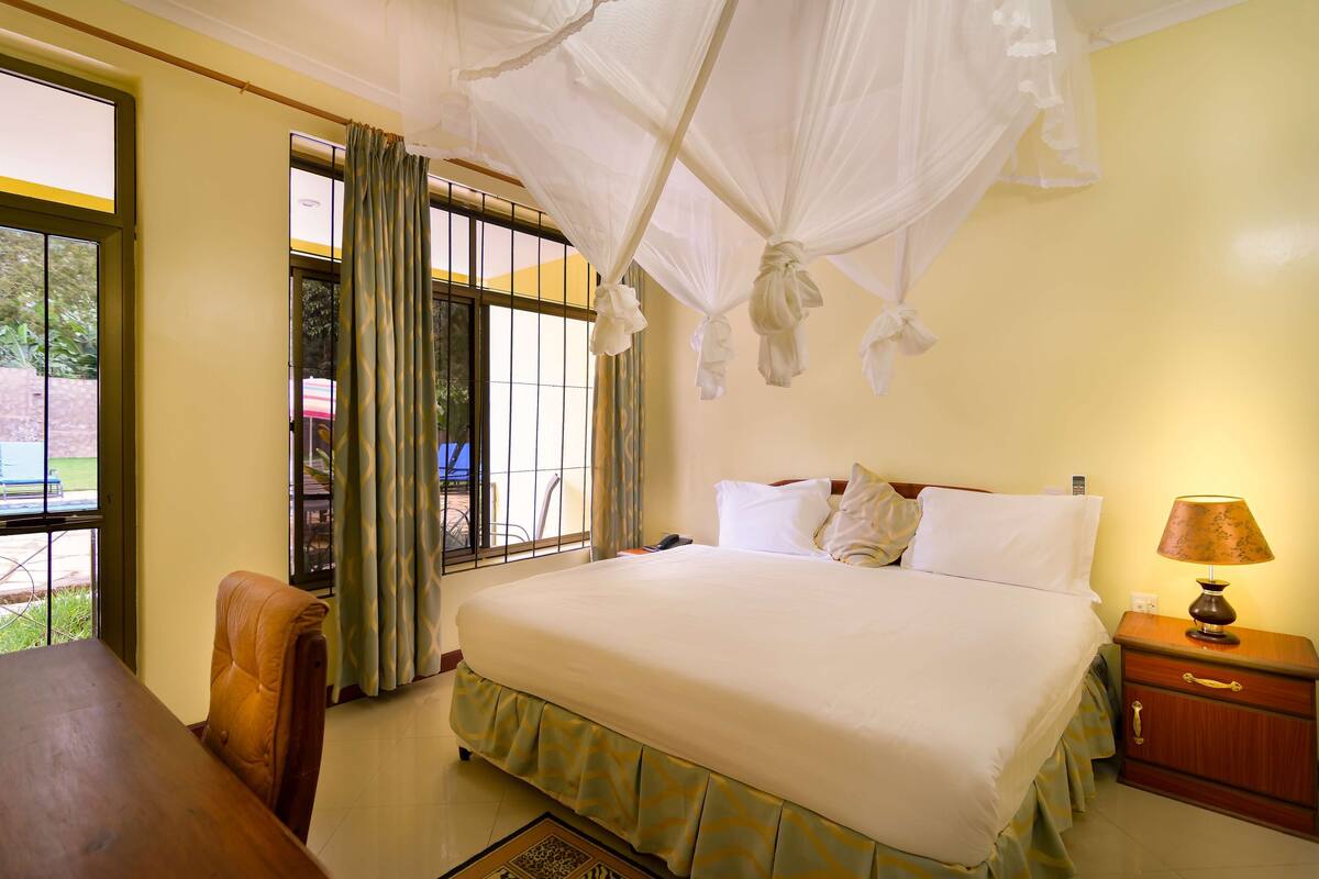 Una habitación de hotel con una cama grande, una mesita de noche de madera, una mesa y una ventana con cortinas.