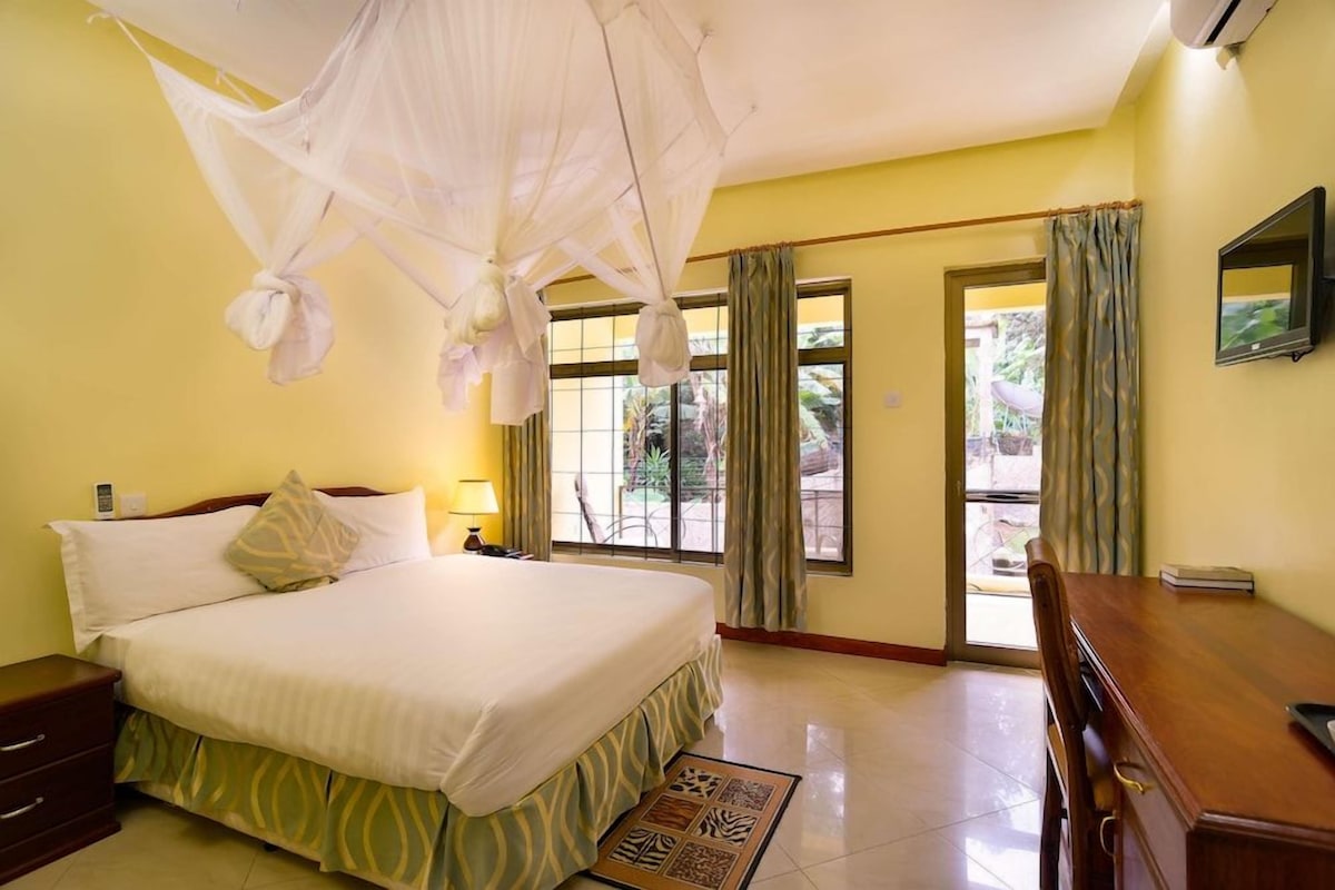 A hotel room with a bed, a desk, a chair, a nightstand, a lamp, and a window with curtains.