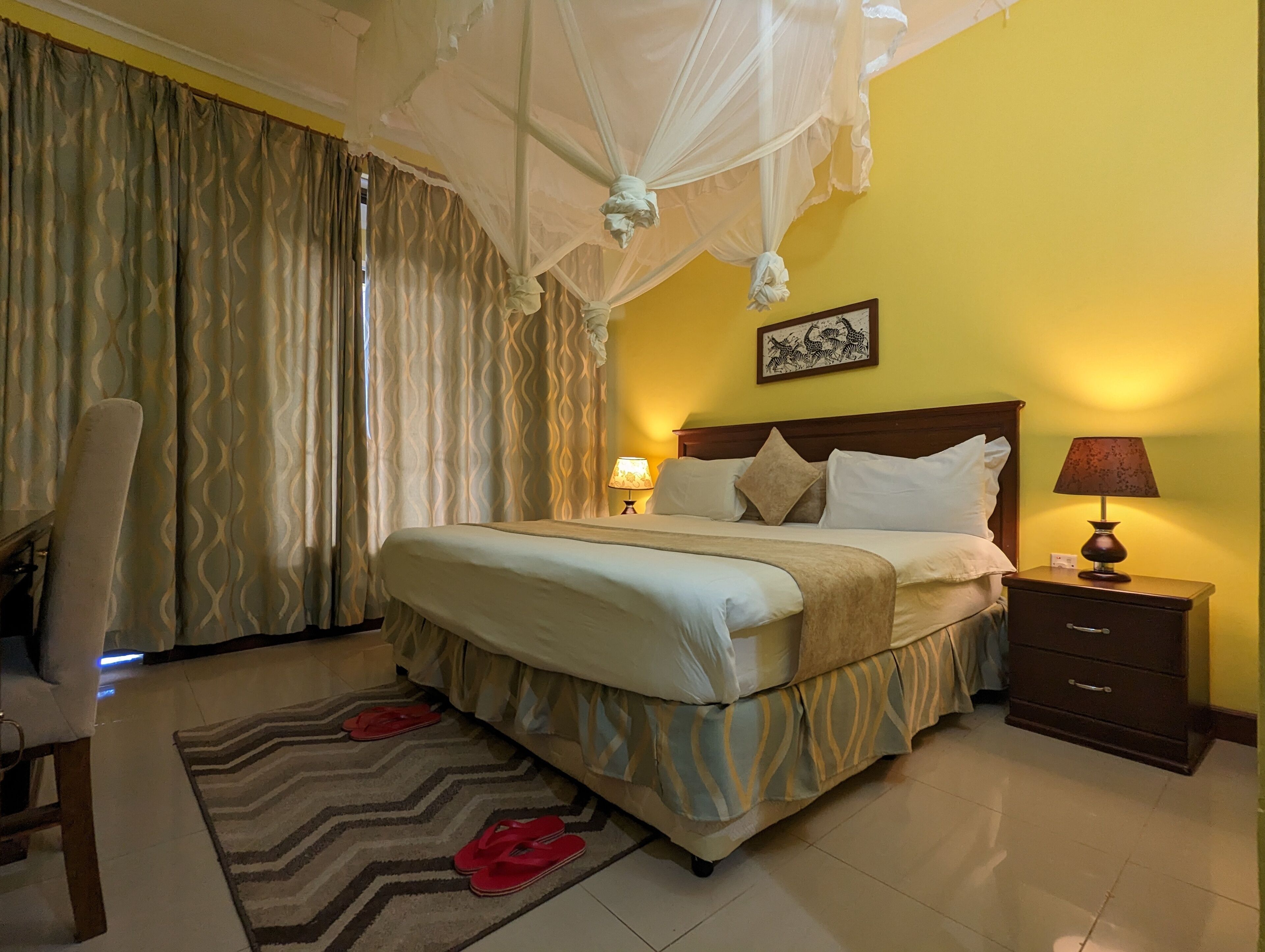 A neatly made bed with a canopy, a bedside table with a lamp, and a chair with a patterned rug.