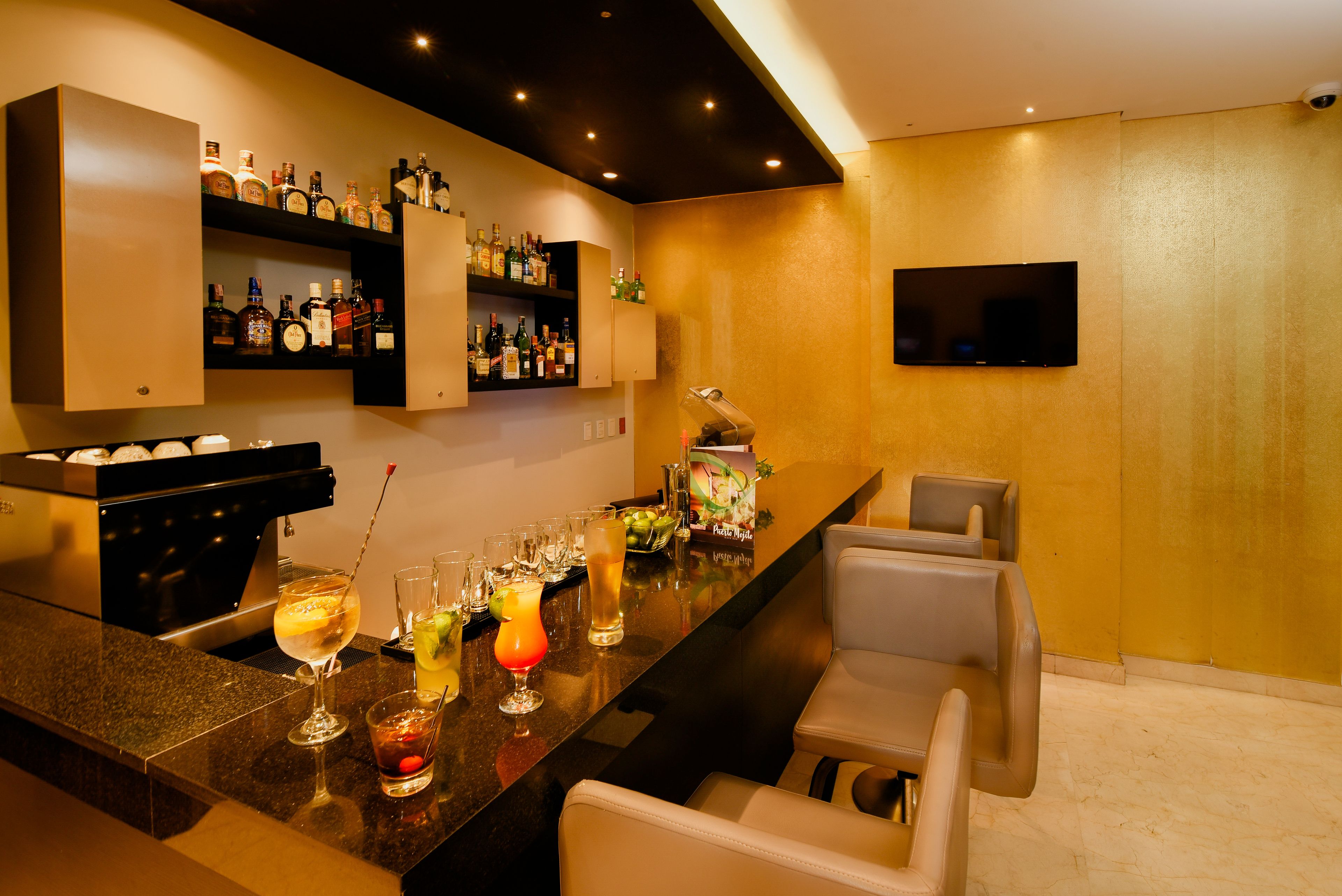 Una zona de bar con una barra alargada, varias estanterías llenas de botellas de licores, taburetes y un televisor de pantalla plana montado en la pared.
