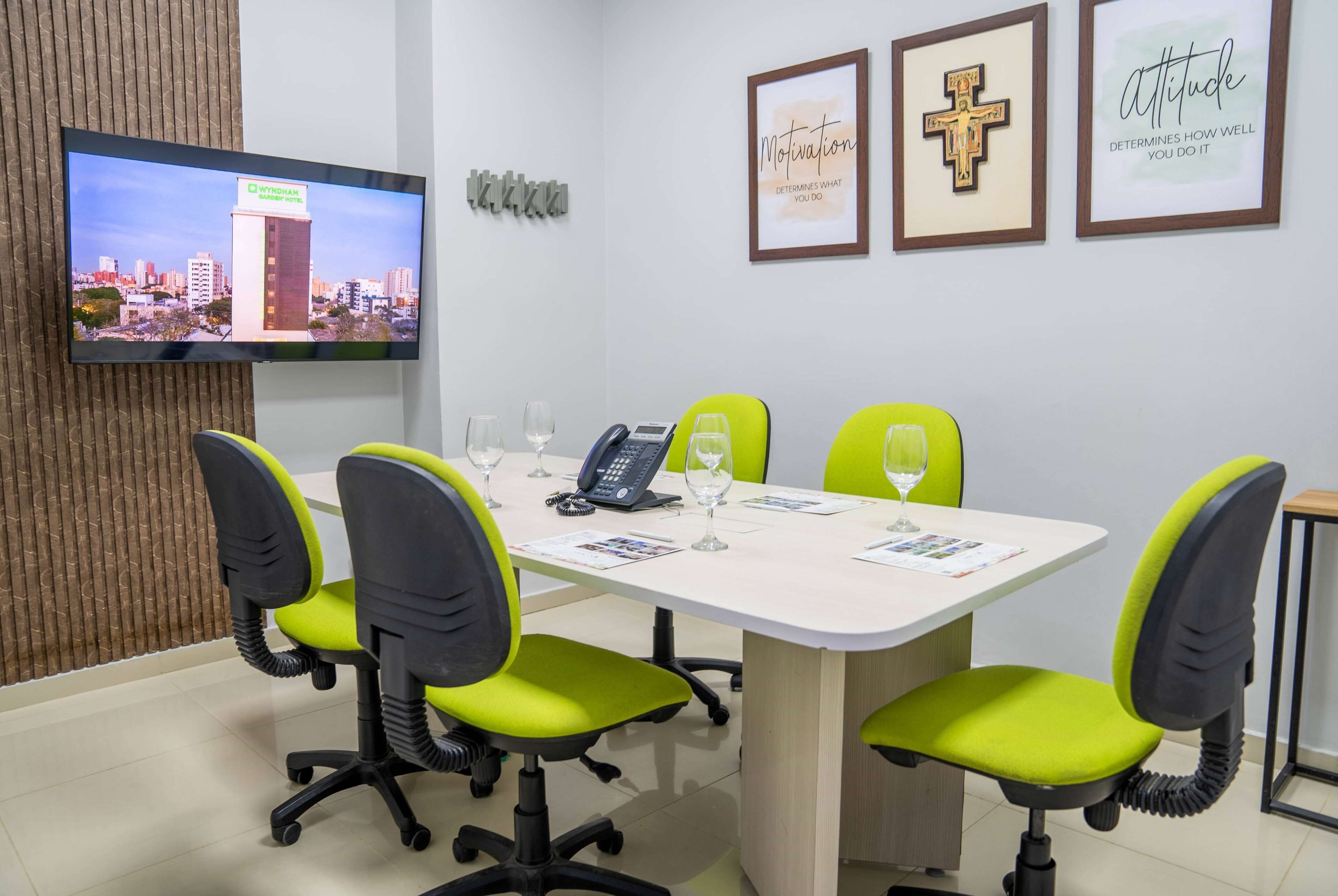 A conference room with a large flat-screen TV displaying a cityscape, a white table, four green and black chairs, a telephone, and three motivational posters on the wall.