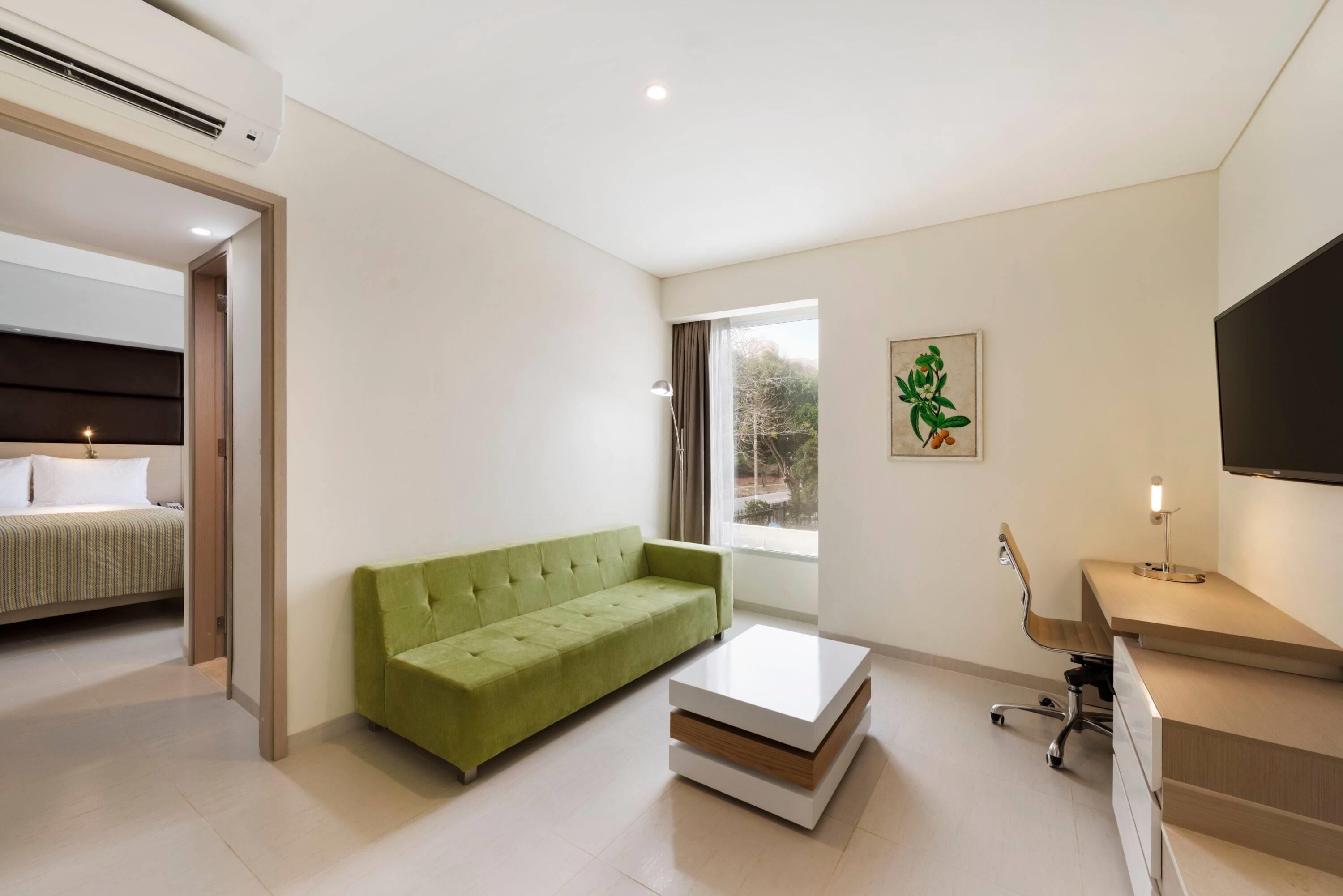 Une chambre d’hôtel moderne avec un canapé vert, une table basse blanche, un bureau avec une lampe et un téléviseur.