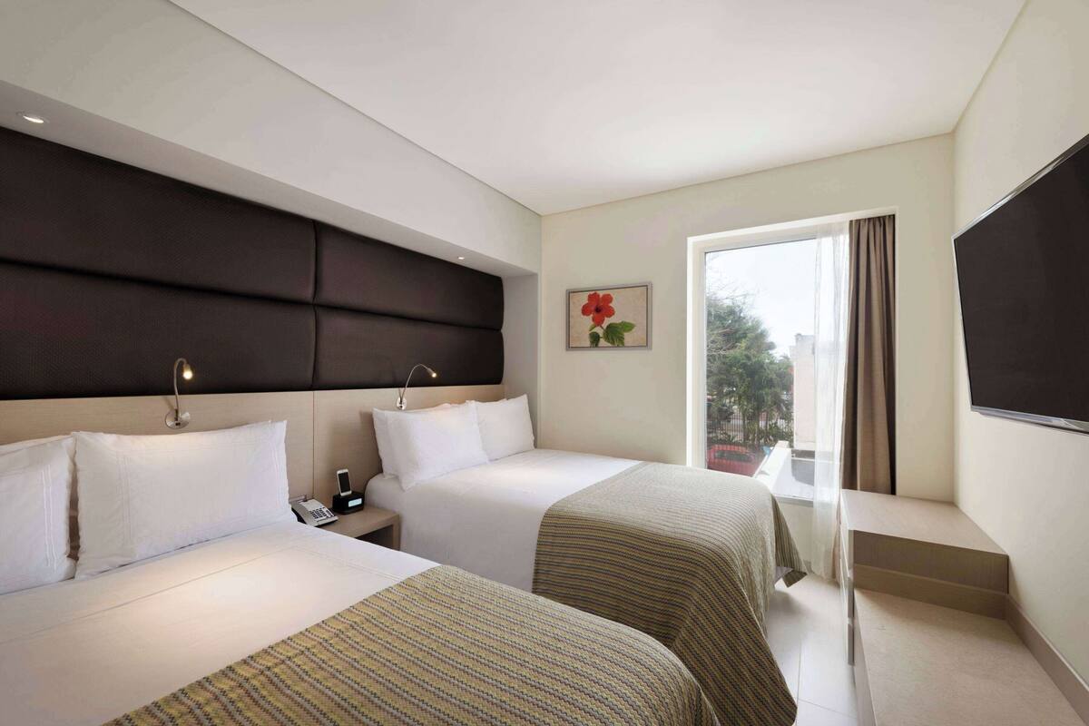 Una habitació d'hotel amb dos llits, un finestral gran amb cortines, un televisor de pantalla plana i una taula petita.