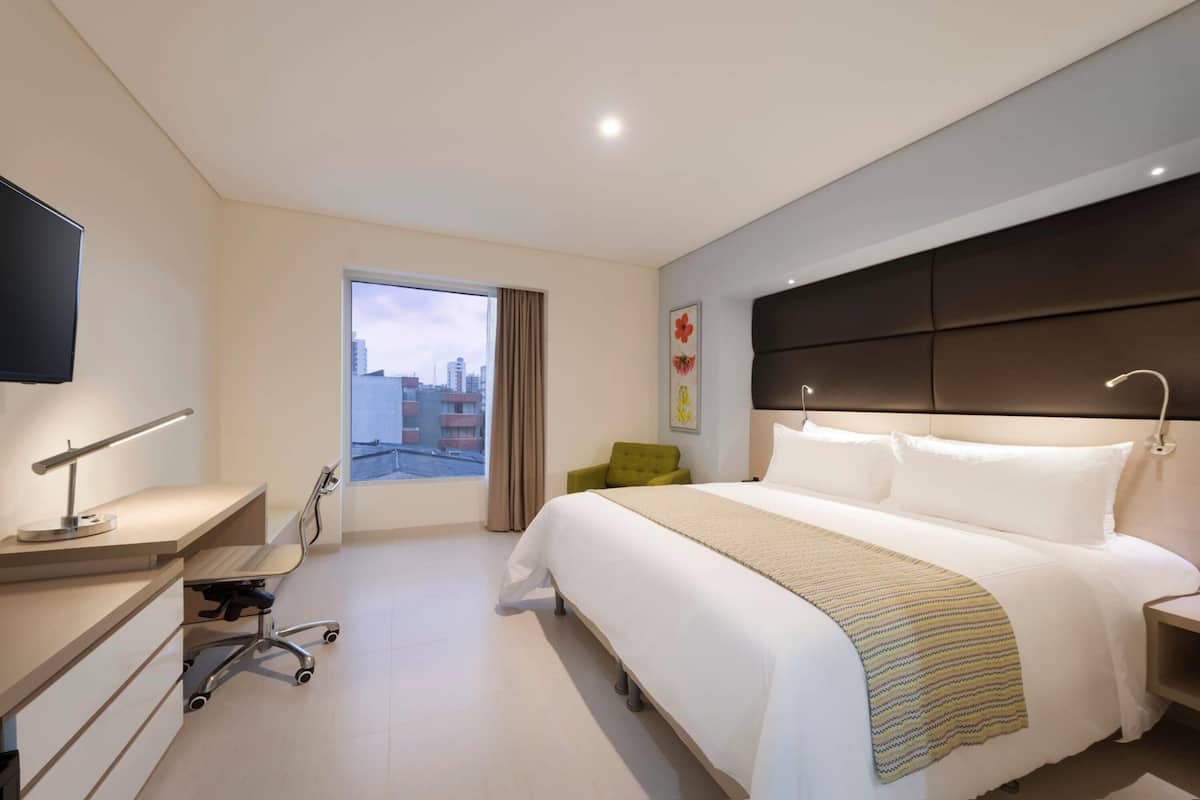Una habitació d'hotel amb un llit gran, una taula de treball amb una làmpada, una cadira i vista a la ciutat per la finestra.