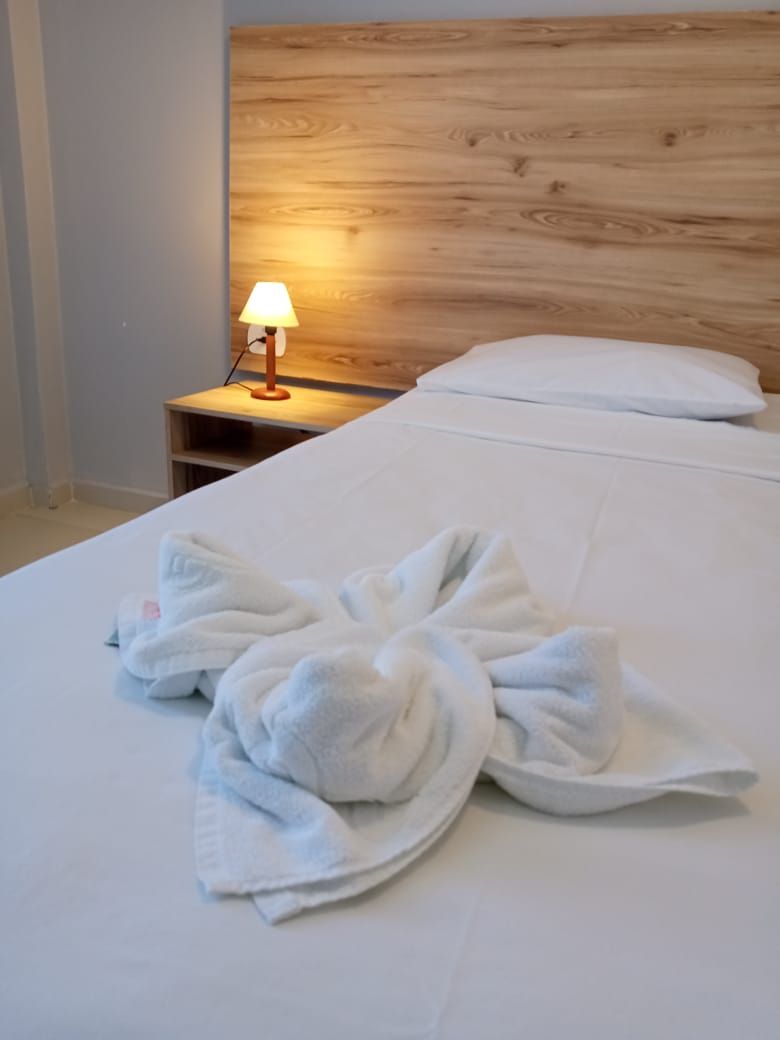 Hotellihuone, jossa on puinen sängynpääty, yöpöytä lampun kanssa sekä valkoisilla vuodevaatteilla pedattu sänky, jonka päällä on pyyhe.