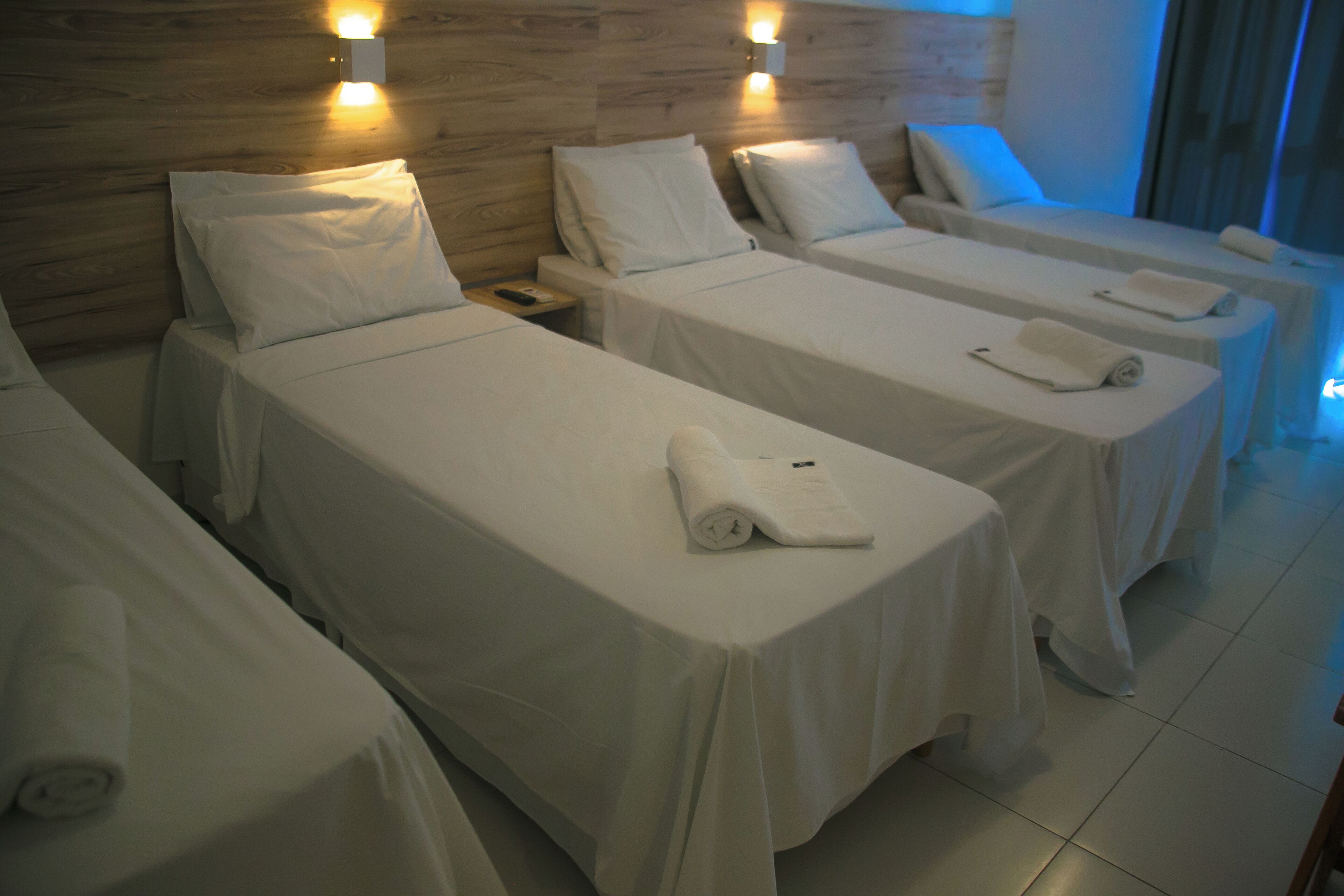 A hotel room with three single beds, each with a white bedspread and pillows, a nightstand with a lamp, and a wooden headboard.