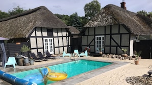 A pool area with a thatched-roof building, lounge chairs, and potted plants. - Farm61 (Tjele)