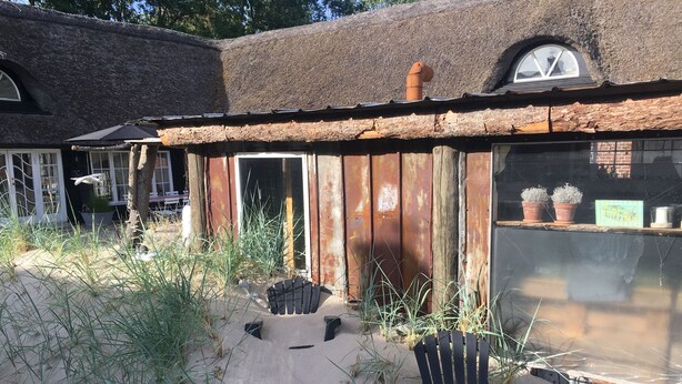 A rustic building with a thatched roof, a wooden door, and a window. There is a black plastic chair and a small black table in the foreground. - Farm61 (Tjele)