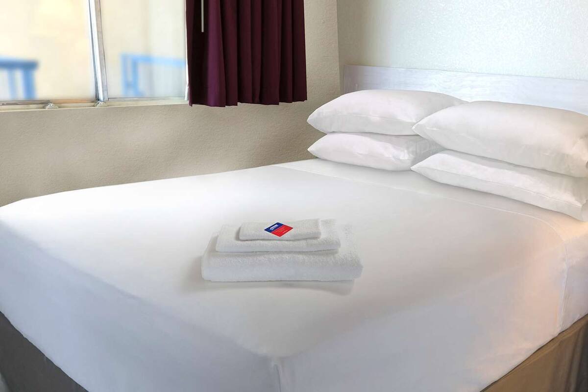 A neatly made bed with white linens and pillows, a folded towel, and a window with curtains.