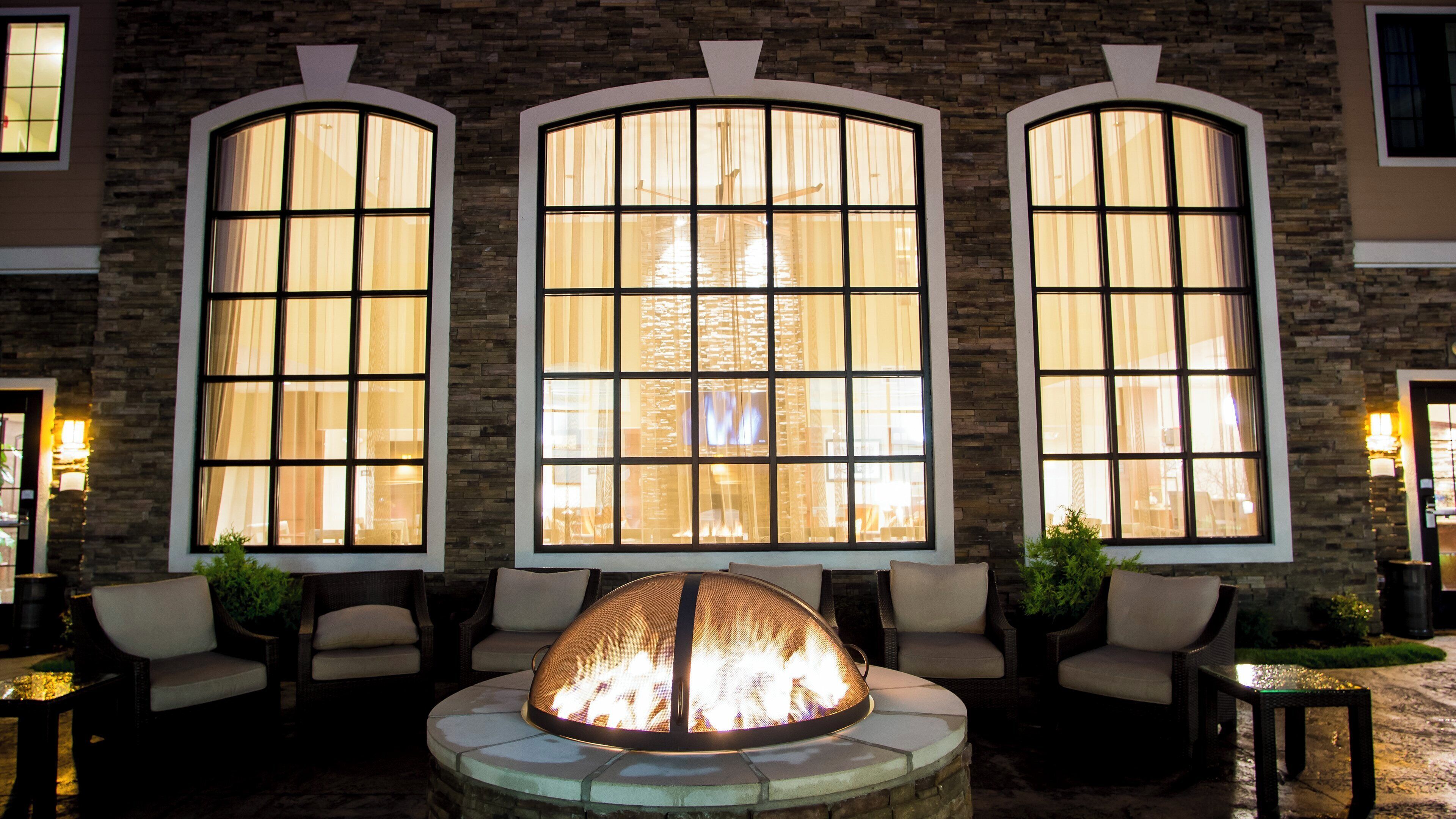A fire pit in the center of a seating area with three large windows on the wall behind.
