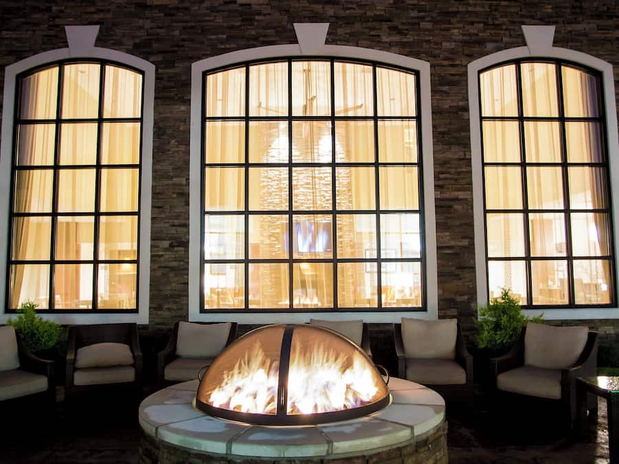 A fire pit in the center of a seating area with three large windows on the wall behind.