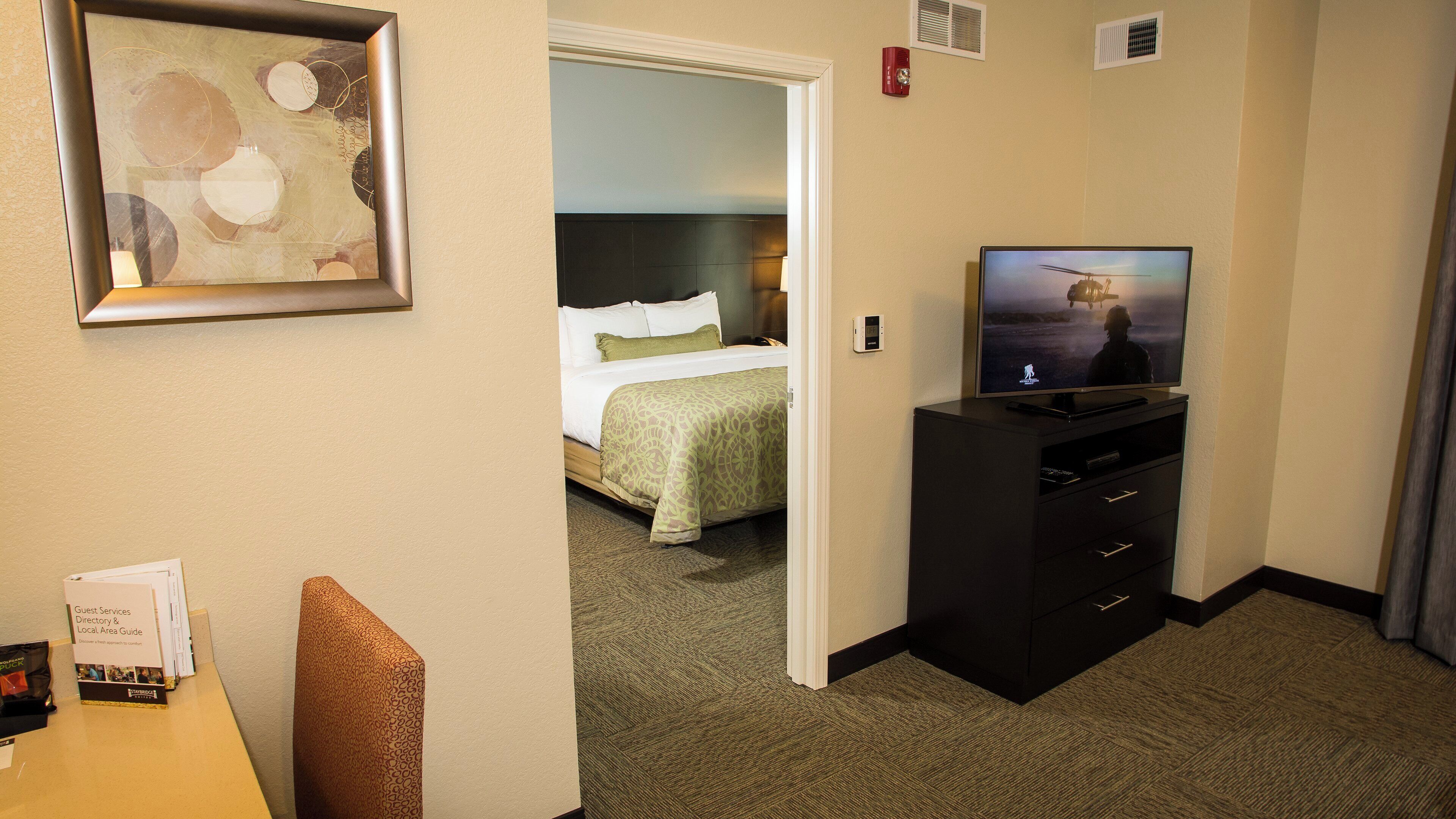 Una habitación de hotel con cama, televisión, escritorio con computadora y un cuadro enmarcado en la pared.