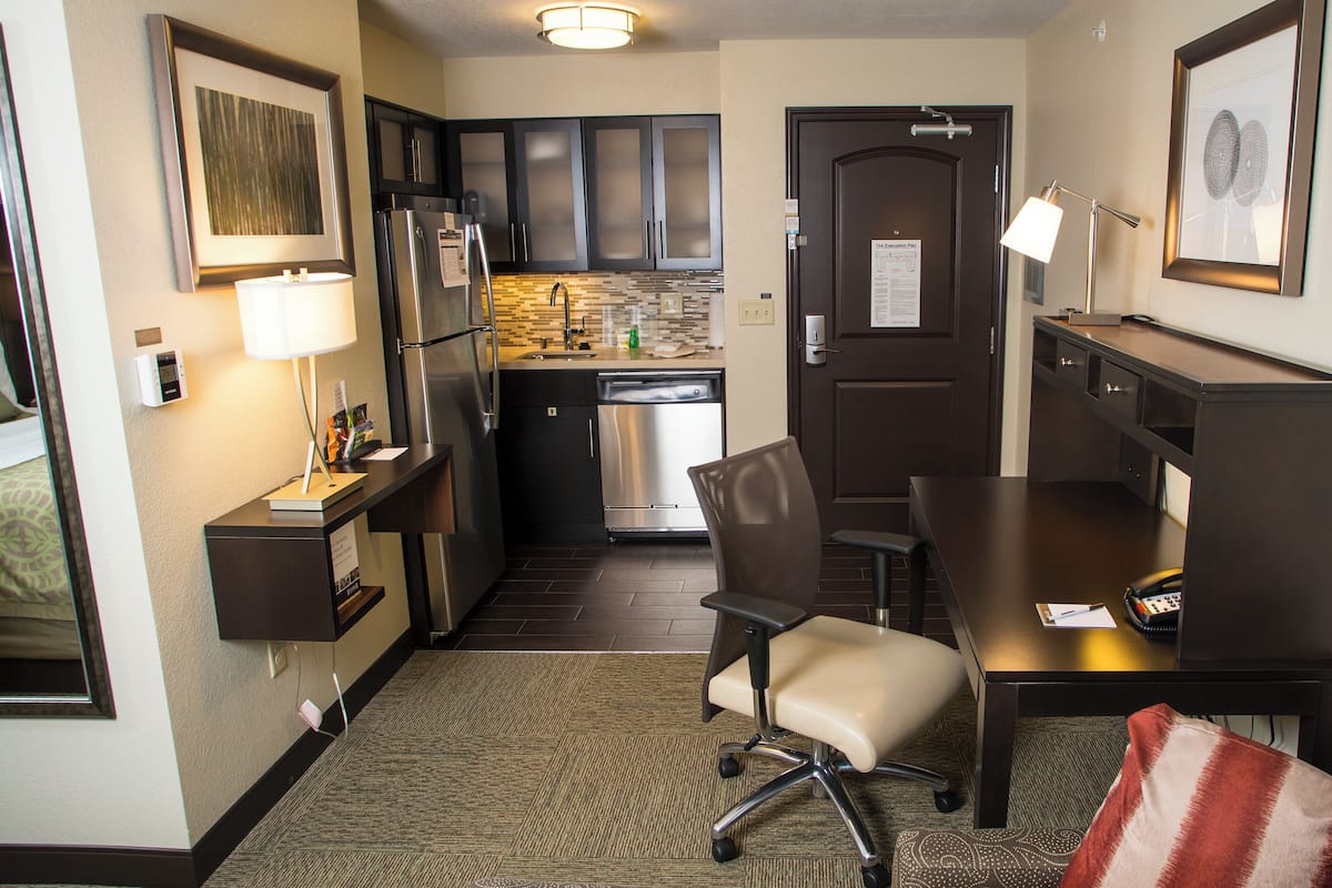 A compact hotel room with a kitchenette, a desk with a chair, a small sofa, and a bedside table.