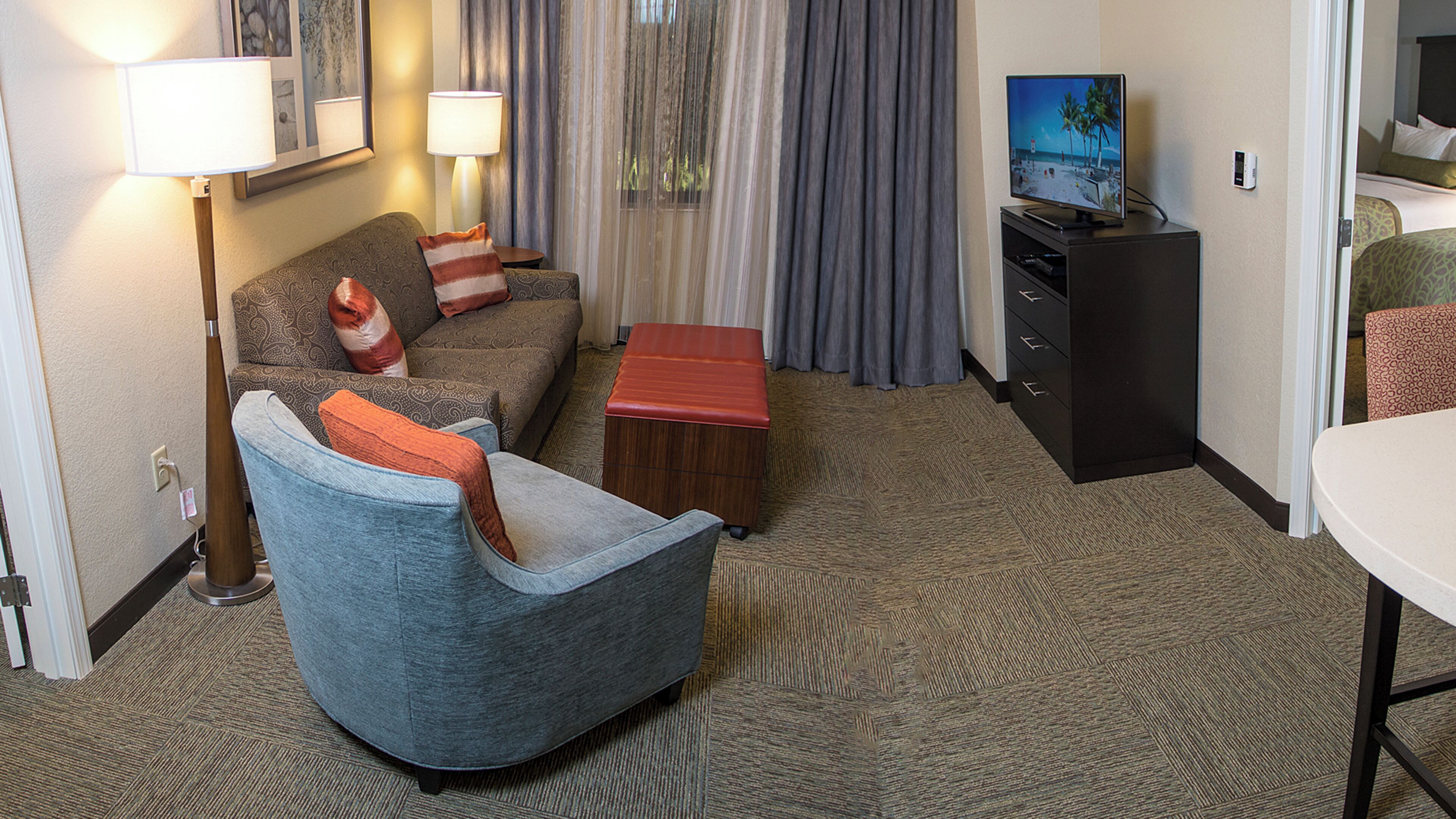 Une chambre d’hôtel avec un fauteuil gris, un canapé agrémenté de coussins à motifs, une table basse en bois, un téléviseur posé sur un meuble, un bureau avec une chaise et une moquette à motifs.