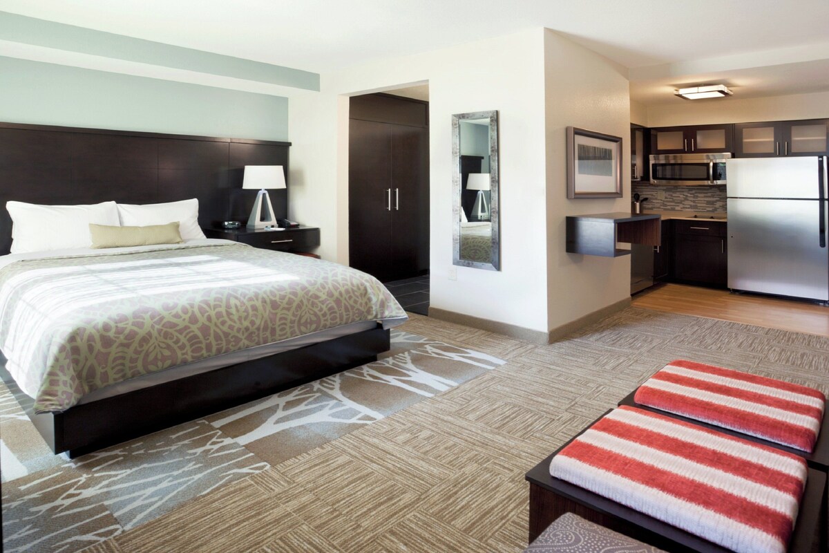 A hotel room with a bed, bedside tables, a nightstand, a bench, a kitchen area with a refrigerator, microwave, and cabinets.