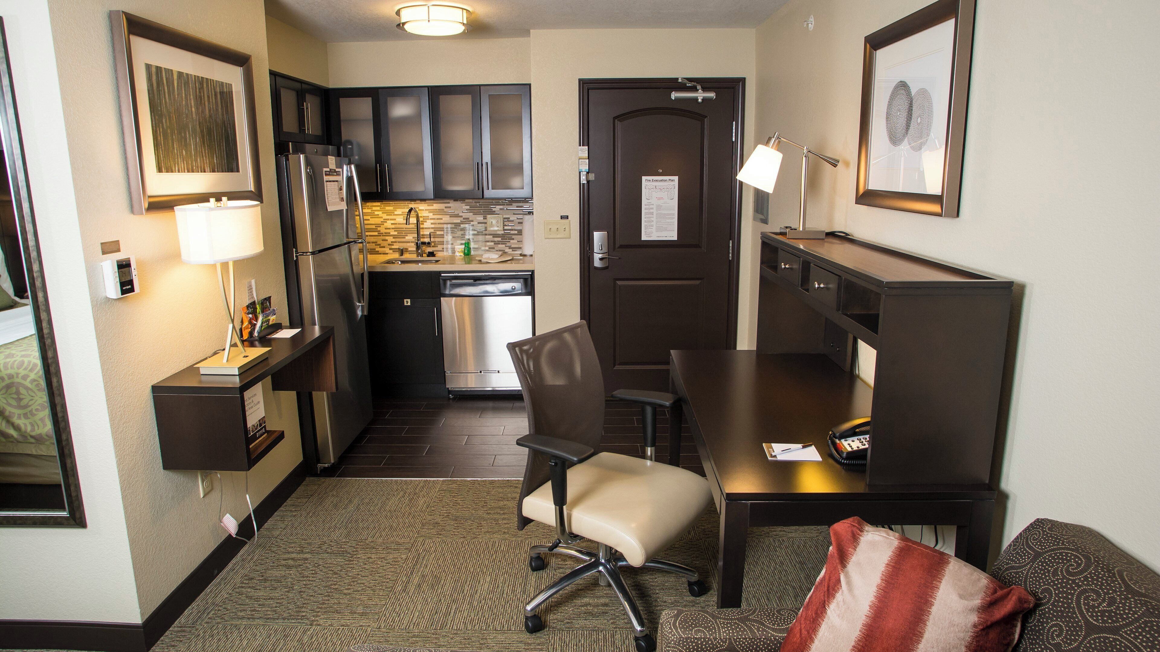 A compact hotel room with a kitchenette, a desk with a chair, a bed, and a small sitting area with a pillow.