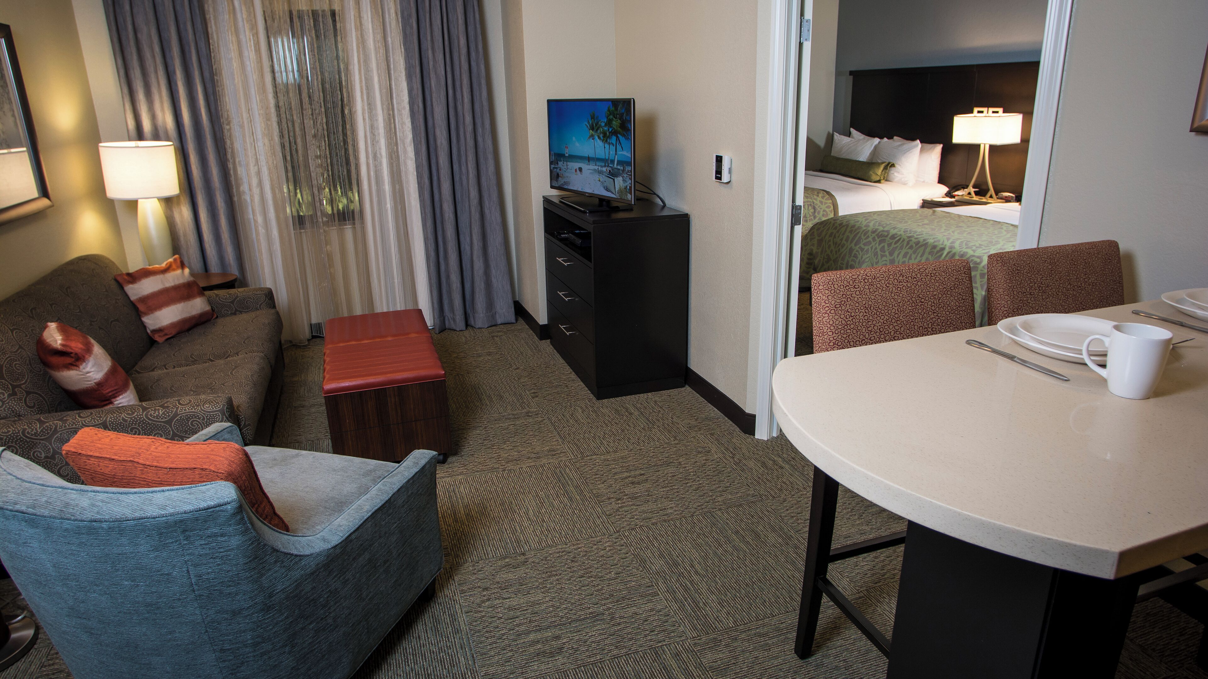 A hotel room with a sofa, armchair, small table, TV, and a bed with a headboard.