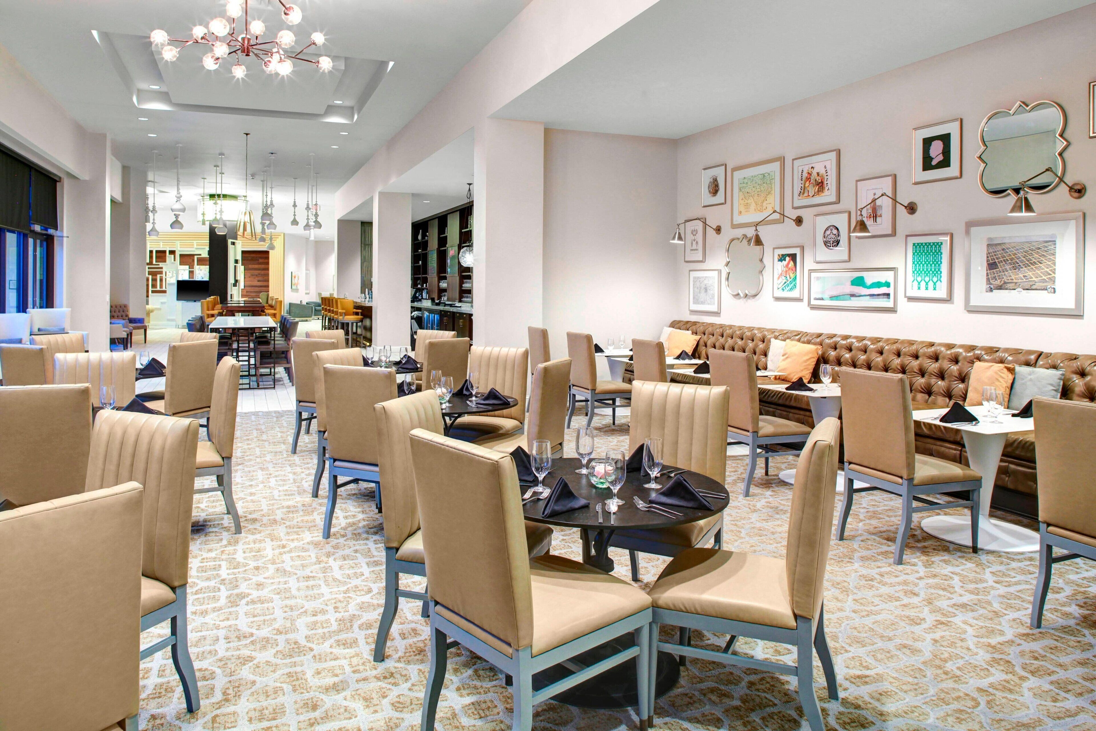 Un comedor amplio con varias mesas y sillas, una zona de estar central con un banco acolchado, y una pared decorada con obras de arte enmarcadas.