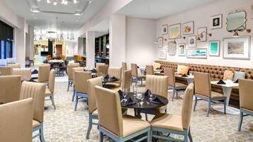 Un comedor amplio con varias mesas y sillas, una zona de estar central con un banco acolchado, y una pared decorada con obras de arte enmarcadas.
