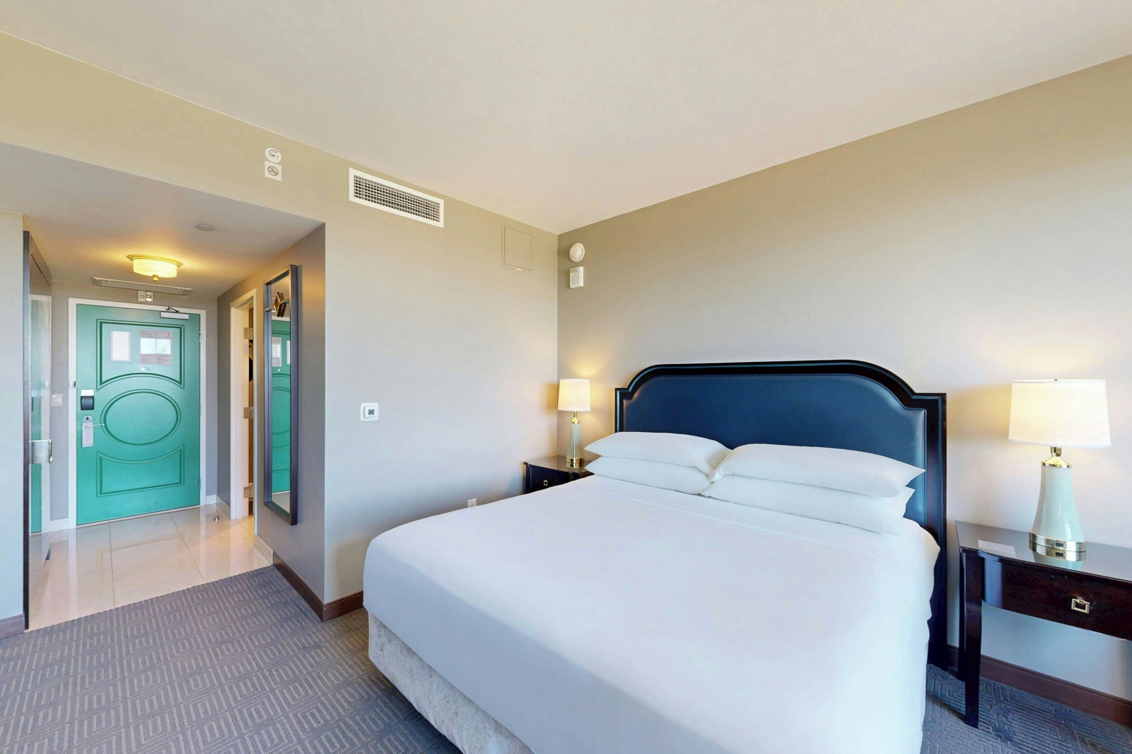 A hotel room with a large bed, two bedside tables with lamps, a door, and a wall-mounted light fixture.
