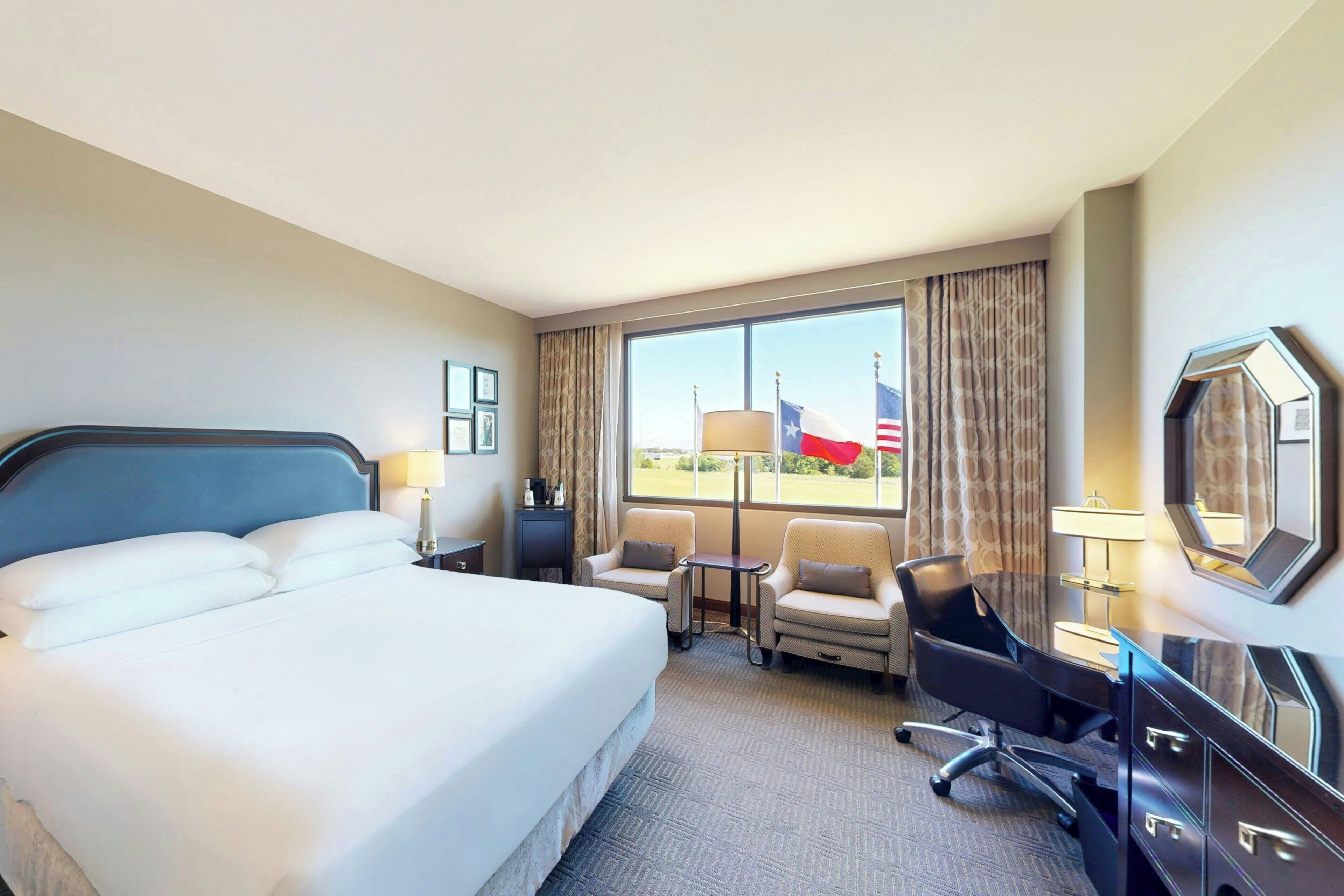 A hotel room with a large bed, two armchairs, a desk, and a view of a flag outside the window.